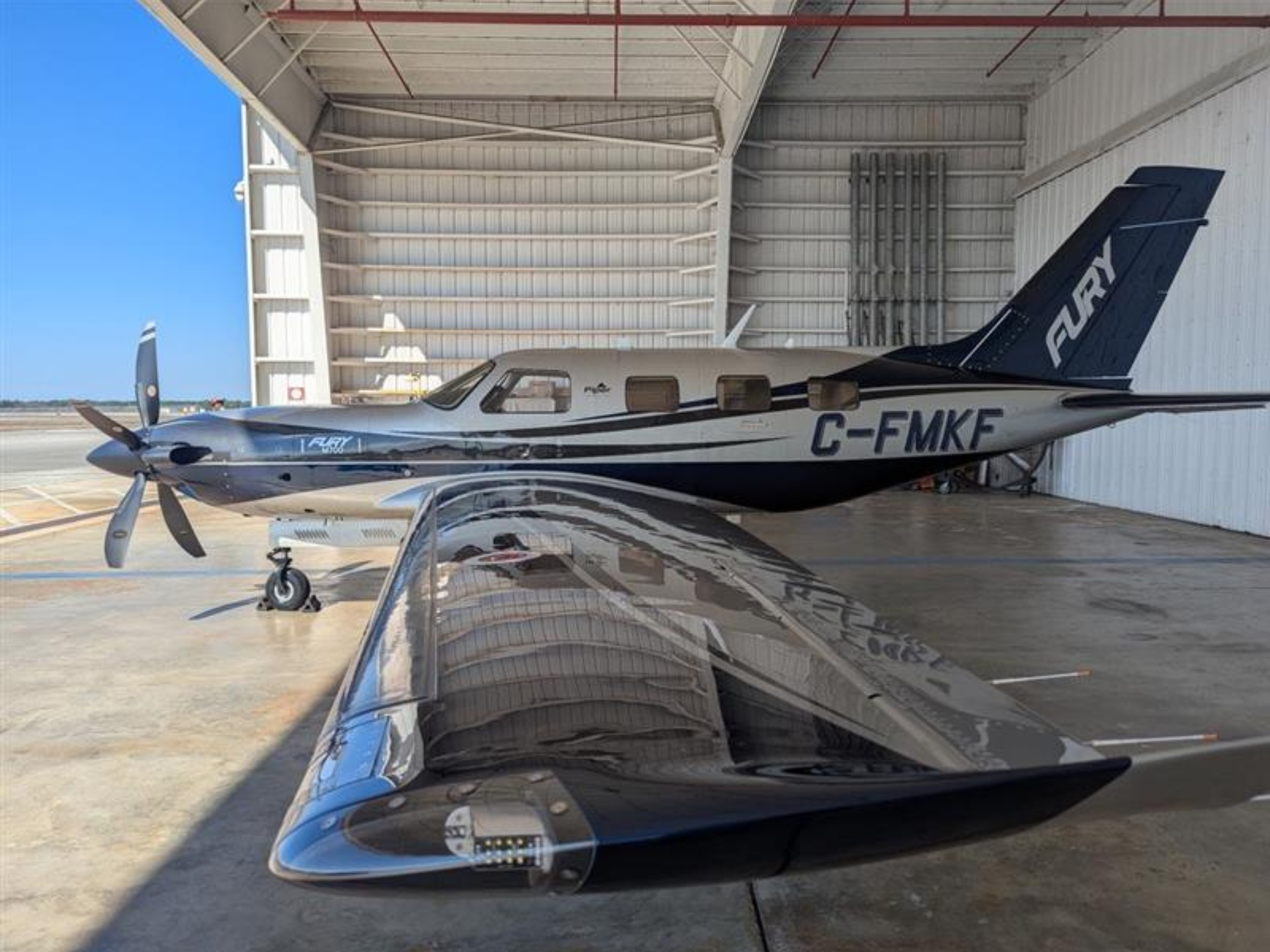 Single-engine propeller airplane parked inside a hangar, silver fuselage with blue accents and registration C-FMKF.