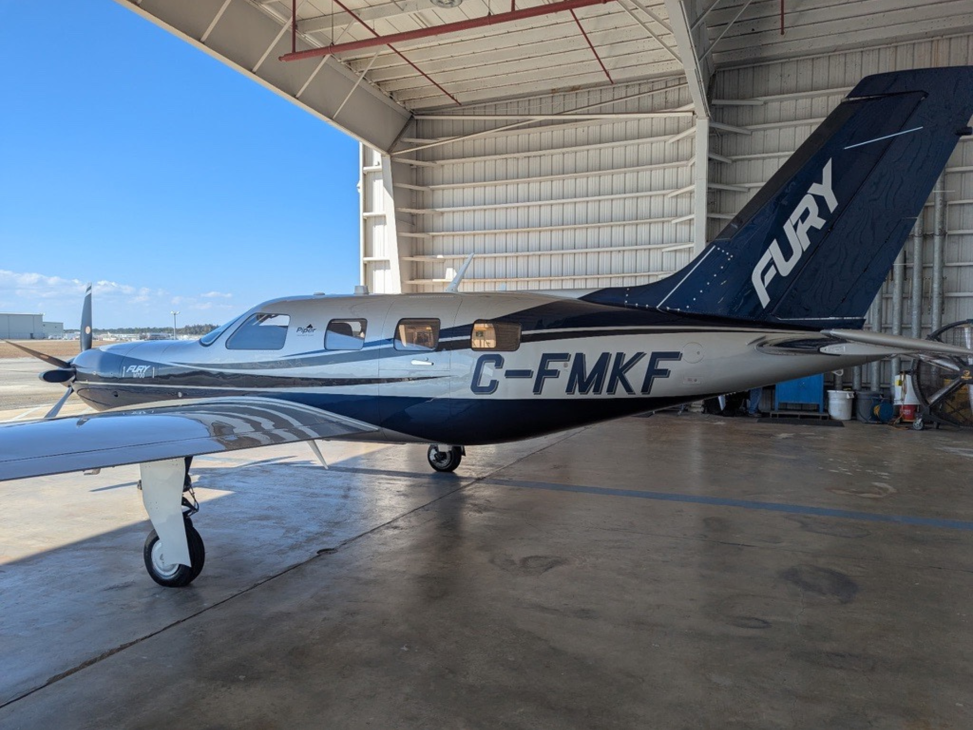 Single-engine light aircraft parked inside a hangar, white and blue livery, registration C-FMKF along the fuselage, blue tail with white lettering visible.