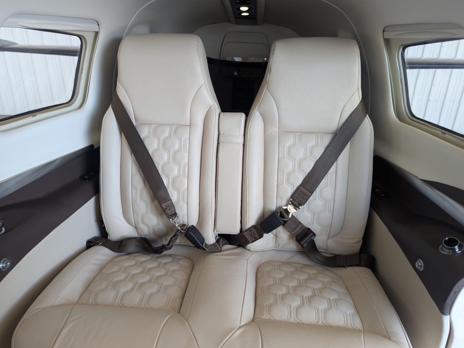Two beige leather airplane seats with buckled seat belts in a narrow cabin interior.