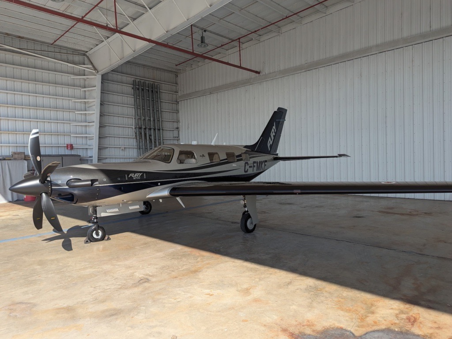 Light single-engine airplane parked in a metal hangar, propeller at the nose and cockpit windows visible.