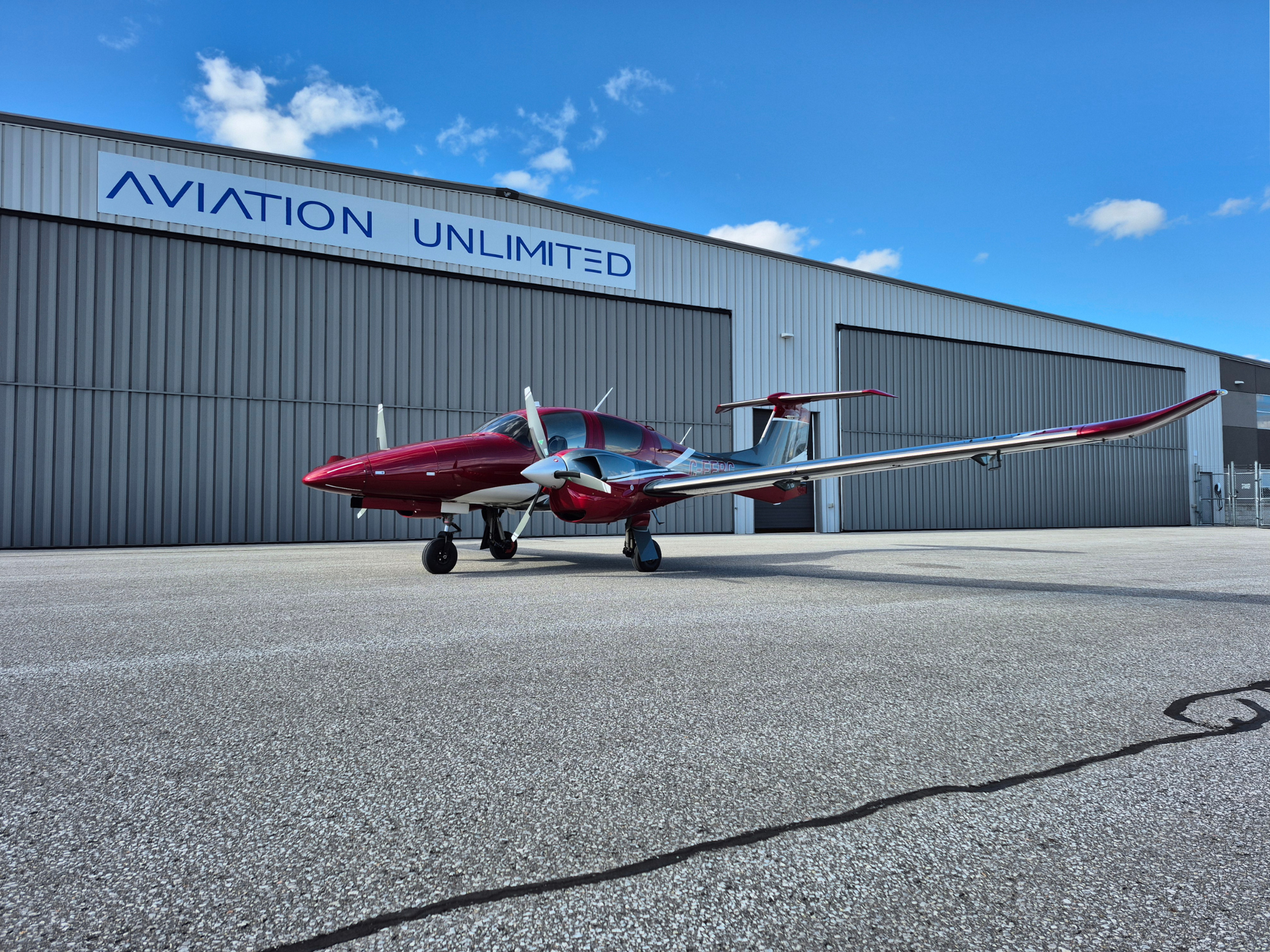 Red and white propeller-powered light aircraft parked on a concrete tarmac in front of a gray hangar reading 'AVIATION UNLIMITED' against a blue sky.
