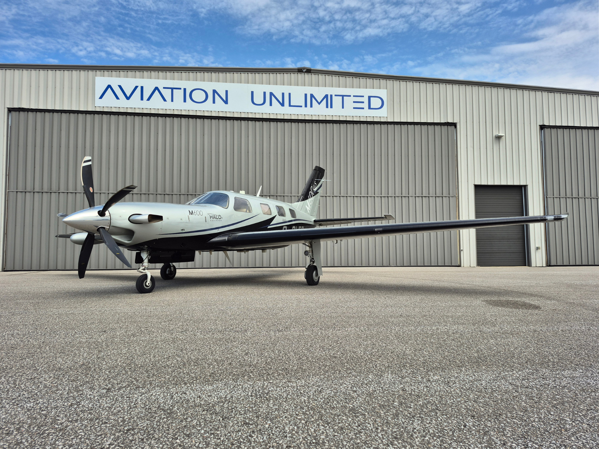 Small silver propeller airplane parked on a tarmac in front of a gray hangar with a blue 'AVIATION UNLIMITED' sign above the doors