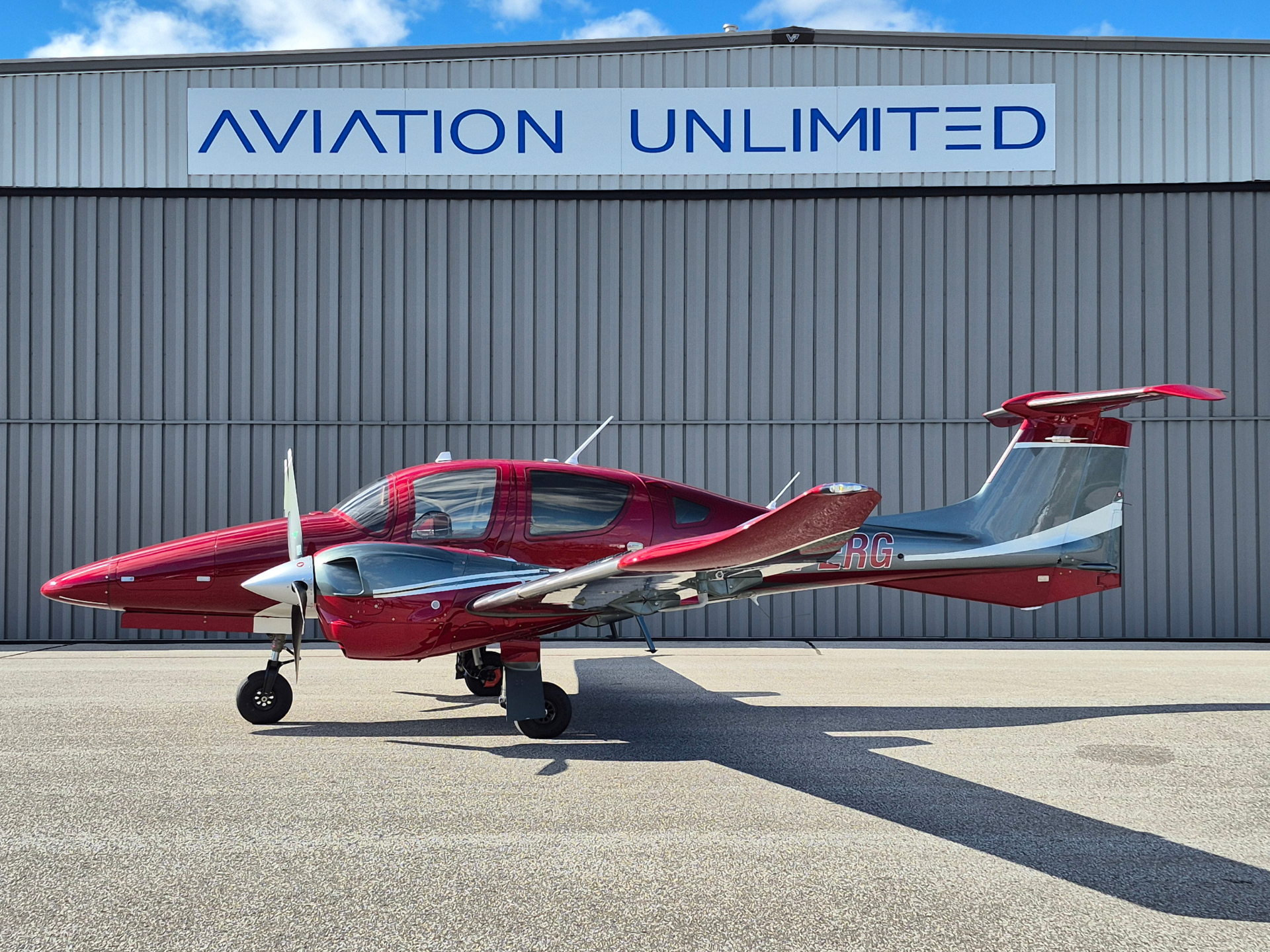 Red small aircraft parked on the tarmac in front of a gray hangar with an 'AVIATION UNLIMITED' sign.