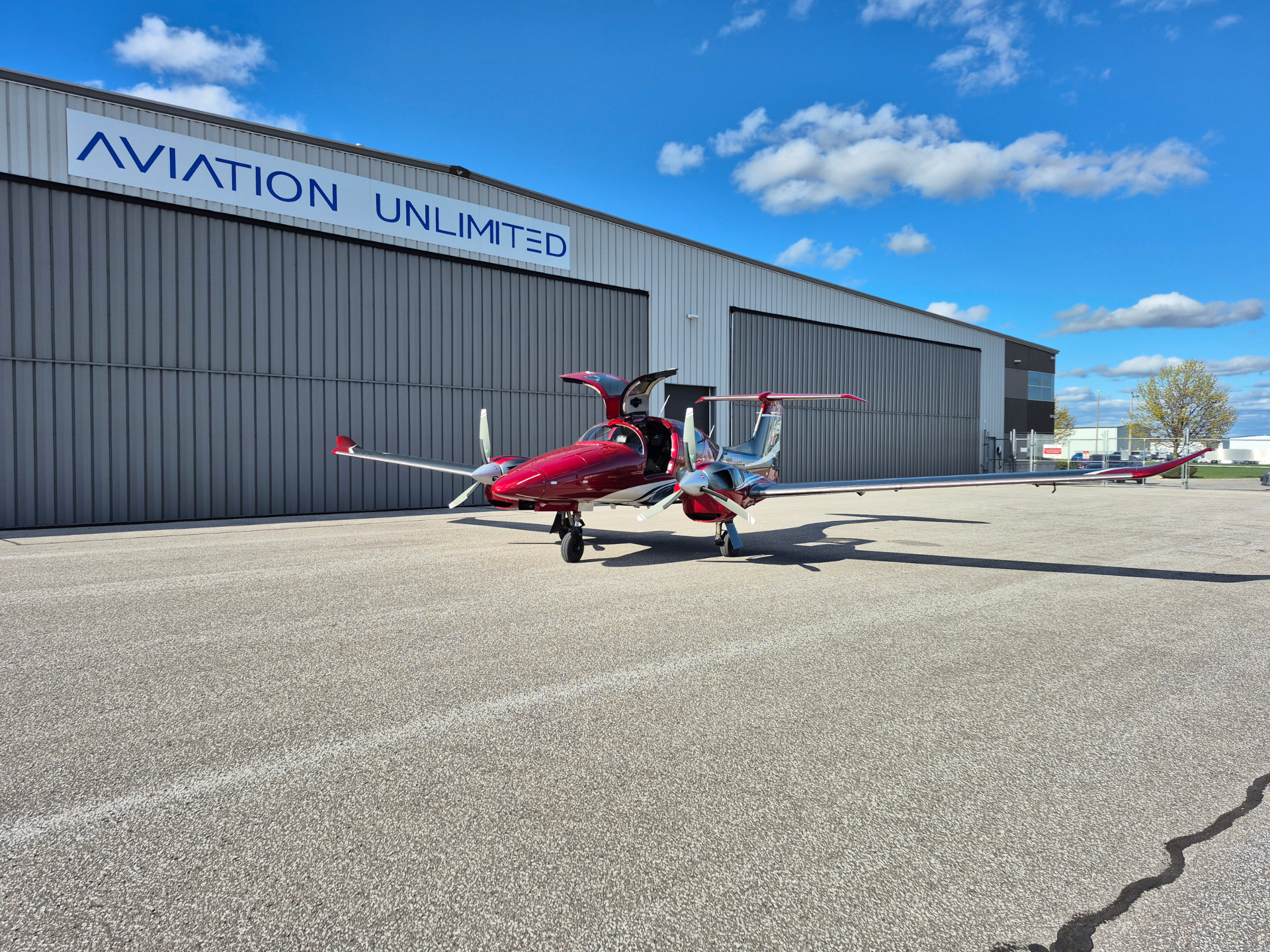 Red twin‑engine airplane with open cockpit doors parked on the tarmac in front of a gray hangar with an 'AVIATION UNLIMITED' sign under a blue sky.