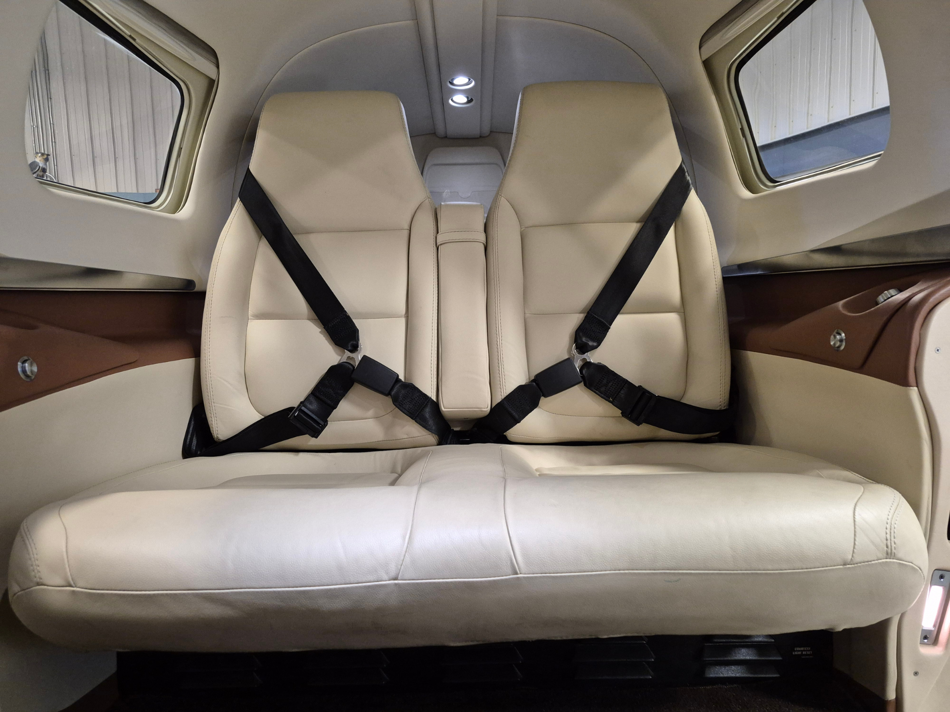 Two beige leather airplane seats with black seat belts facing forward inside a small cabin by windows on either side.