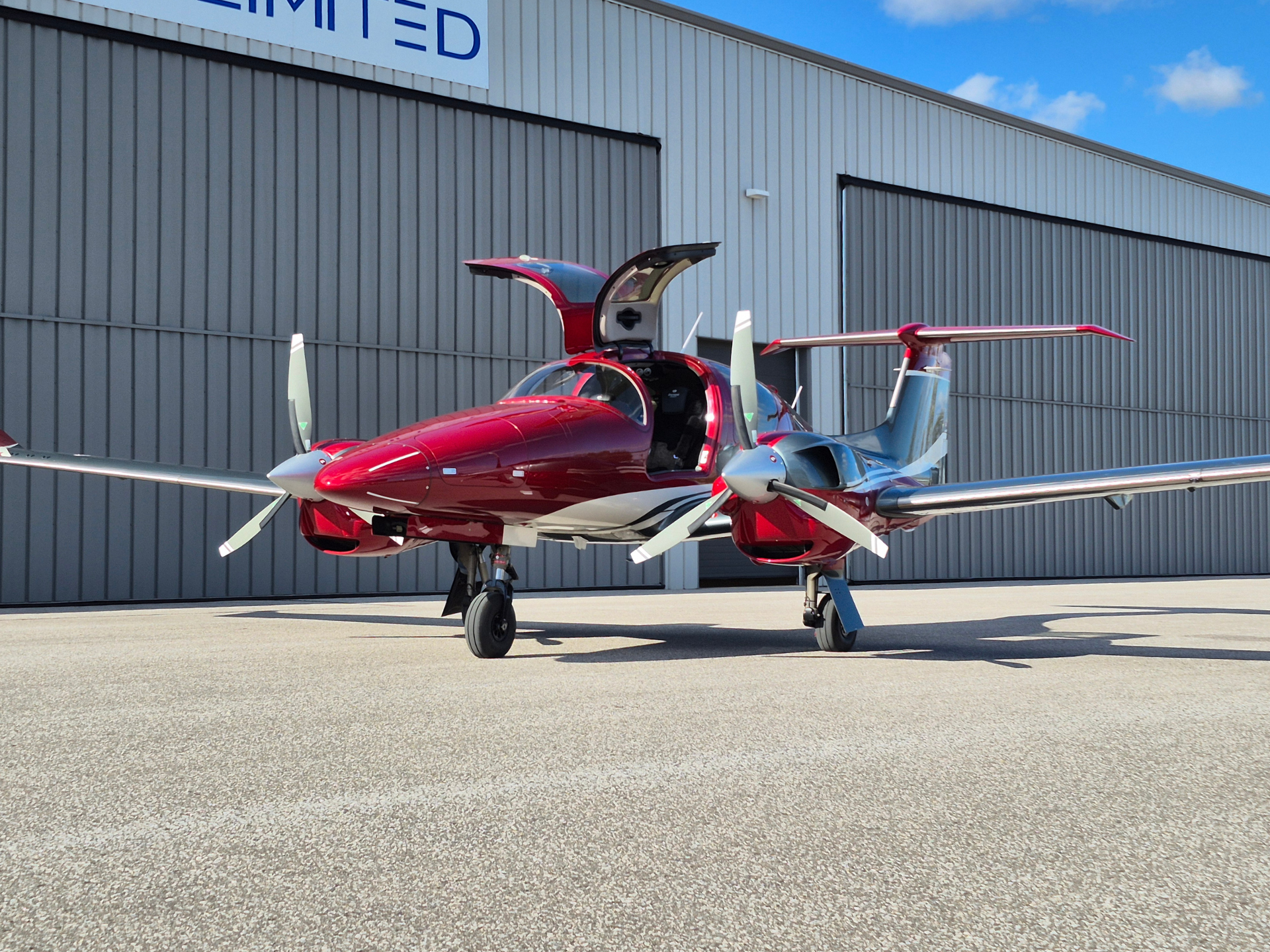 Red twin-engine small airplane with open cockpit doors parked on a tarmac in front of a gray hangar.