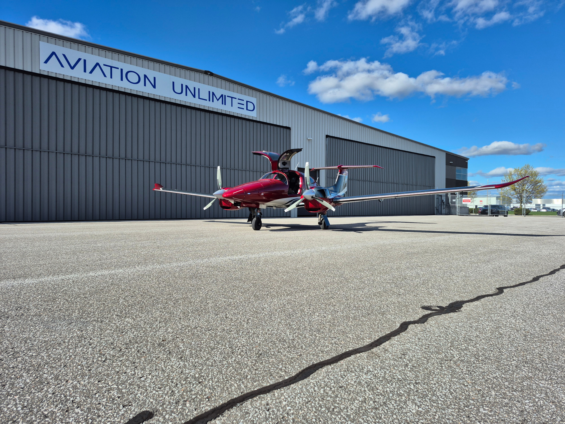 Red and silver twin-engine propeller airplane parked on the tarmac beside a gray aviation hangar under a clear blue sky.