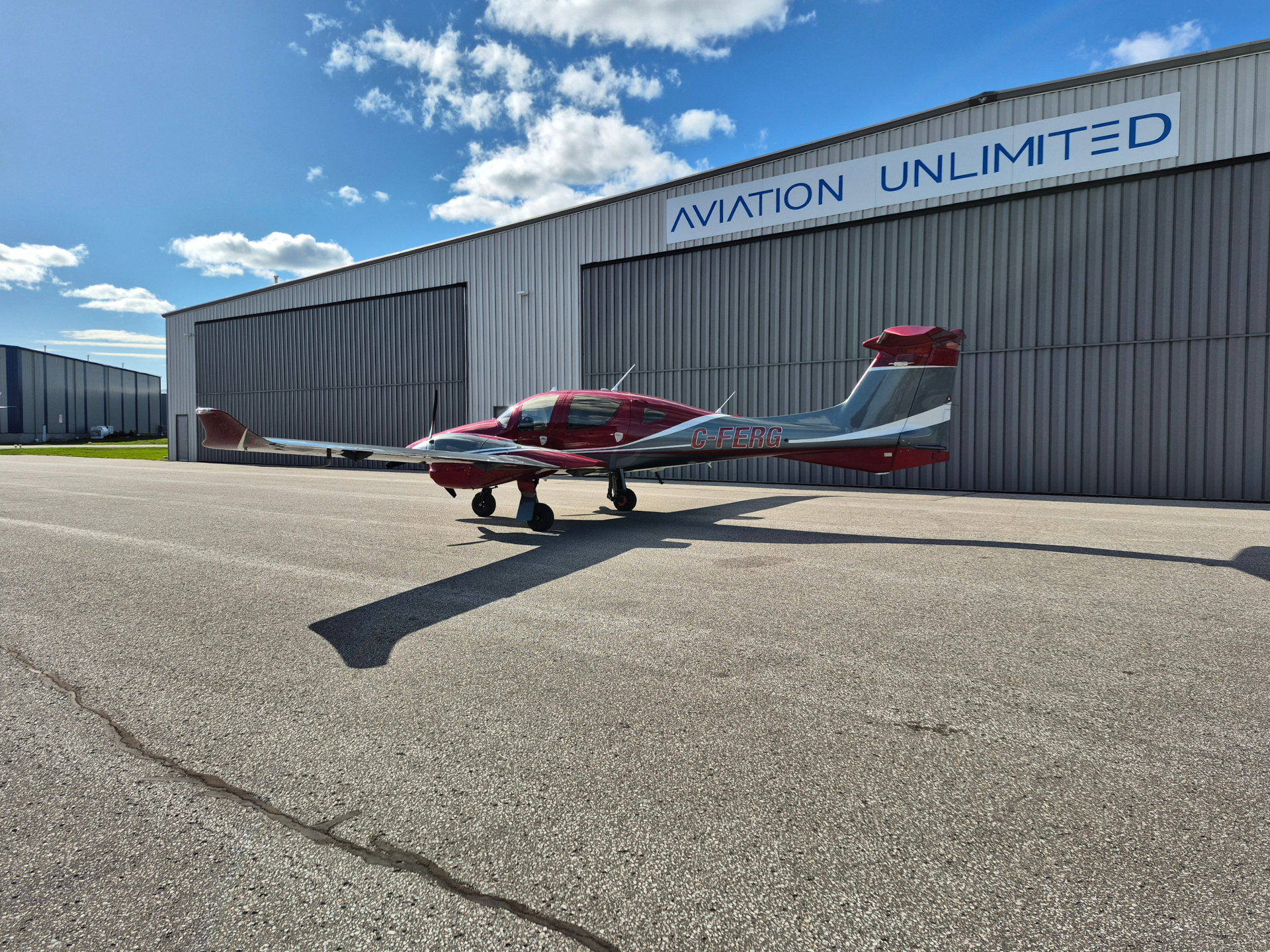 Small red and silver single-engine airplane parked on a tarmac in front of a gray hangar with 'AVIATION UNLIMITED' sign.