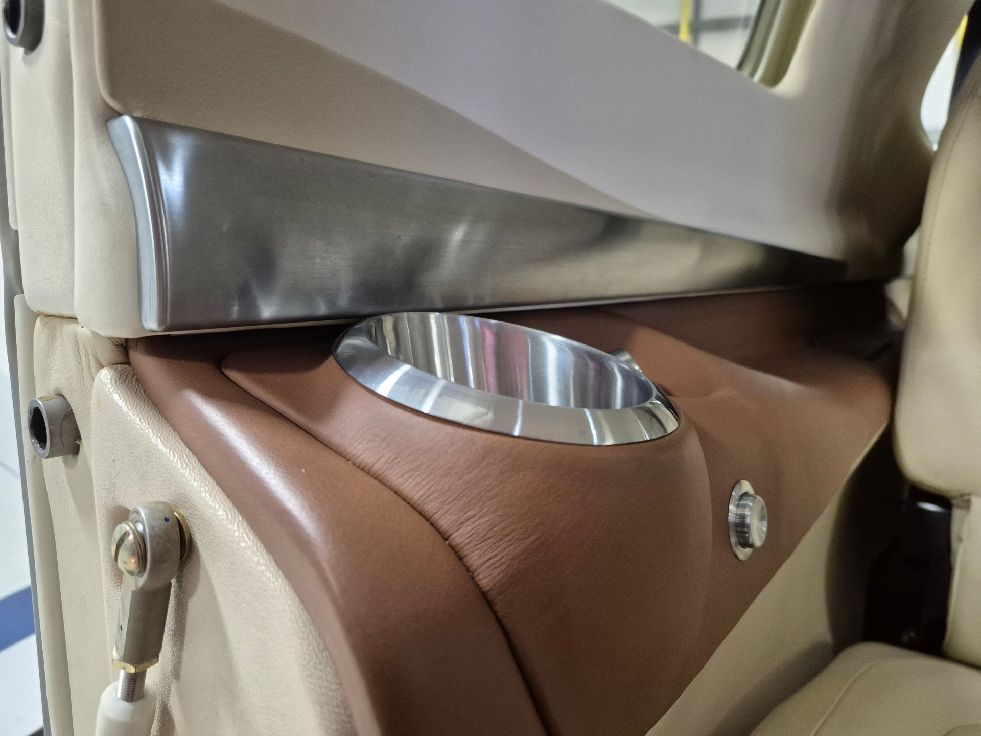 Close-up of a beige car interior with a metallic trim piece and a circular chrome cup holder embedded in brown leather upholstery.
