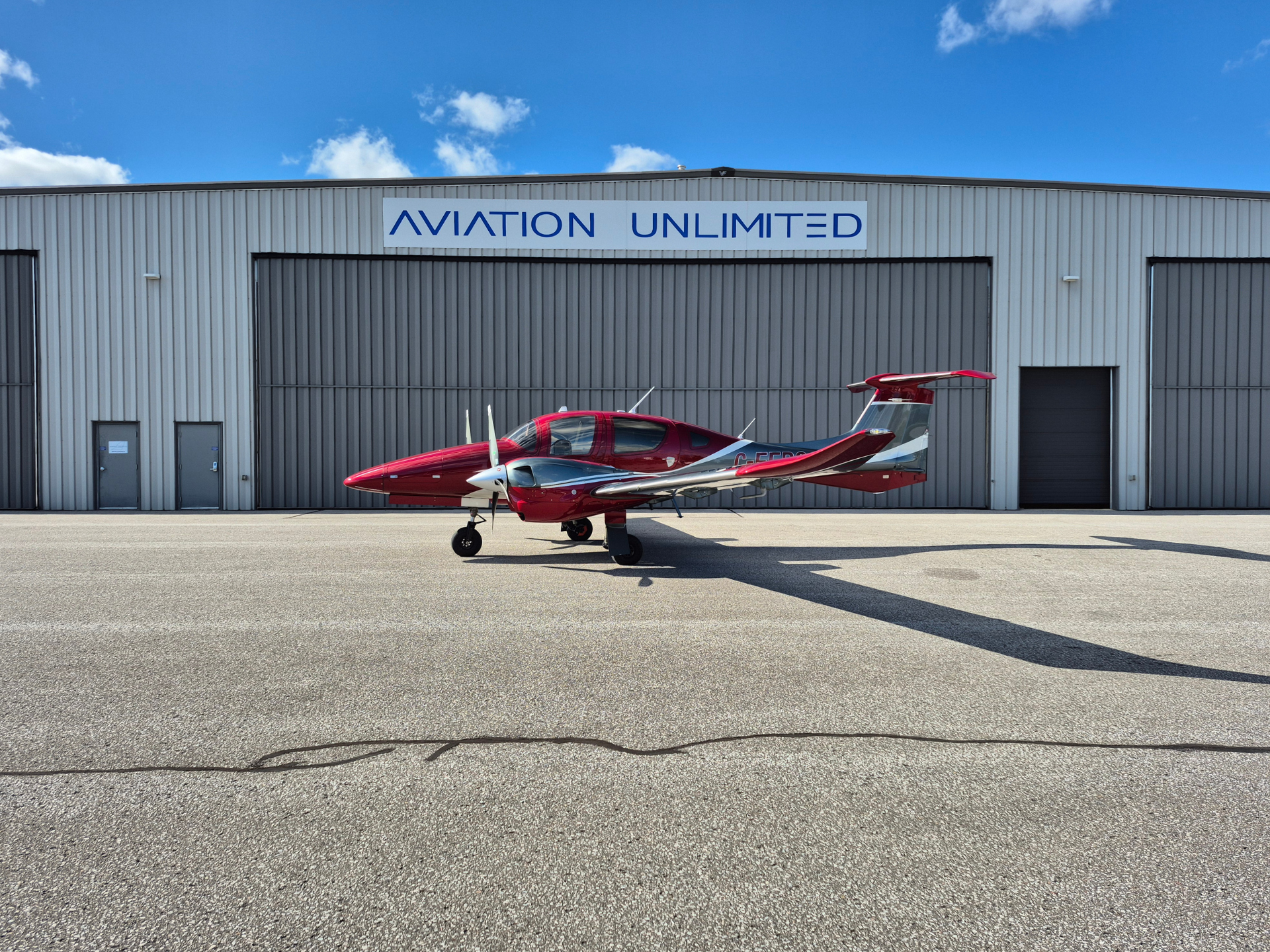 Red single-engine propeller airplane parked on the tarmac in front of a gray hangar labeled 'Aviation Unlimited'.