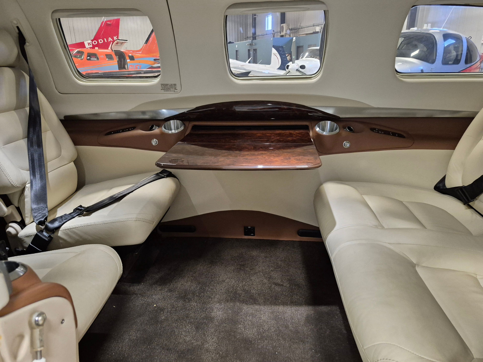 Luxurious private jet cabin with cream leather seats, a polished wood center table, and three windows looking out onto a hangar with parked airplanes.