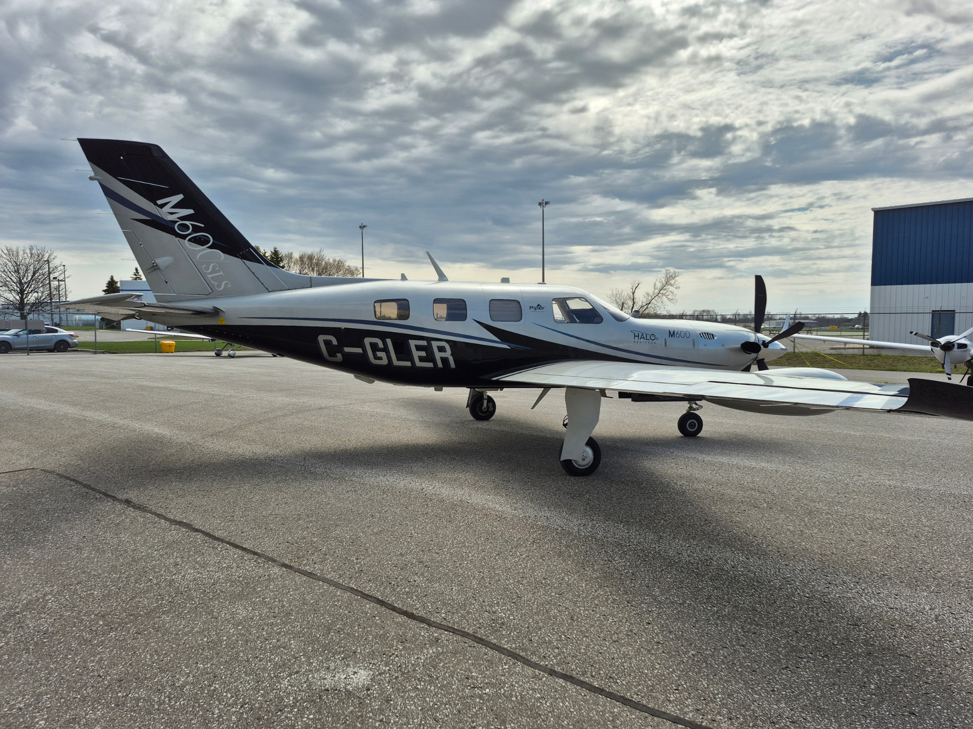 Small twin-prop Piper M600 aircraft parked on a taxiway with hangar in the background, tail number C-GLER and a cloudy sky above.