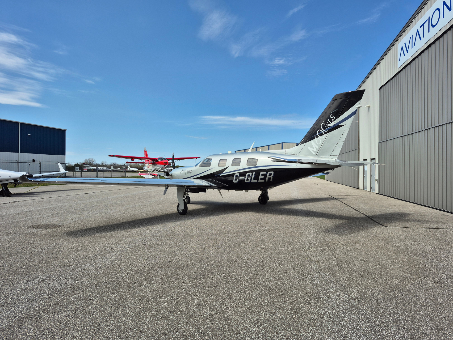 Small silver private plane with C-GLER on the tarmac beside a large hangar under a clear blue sky.