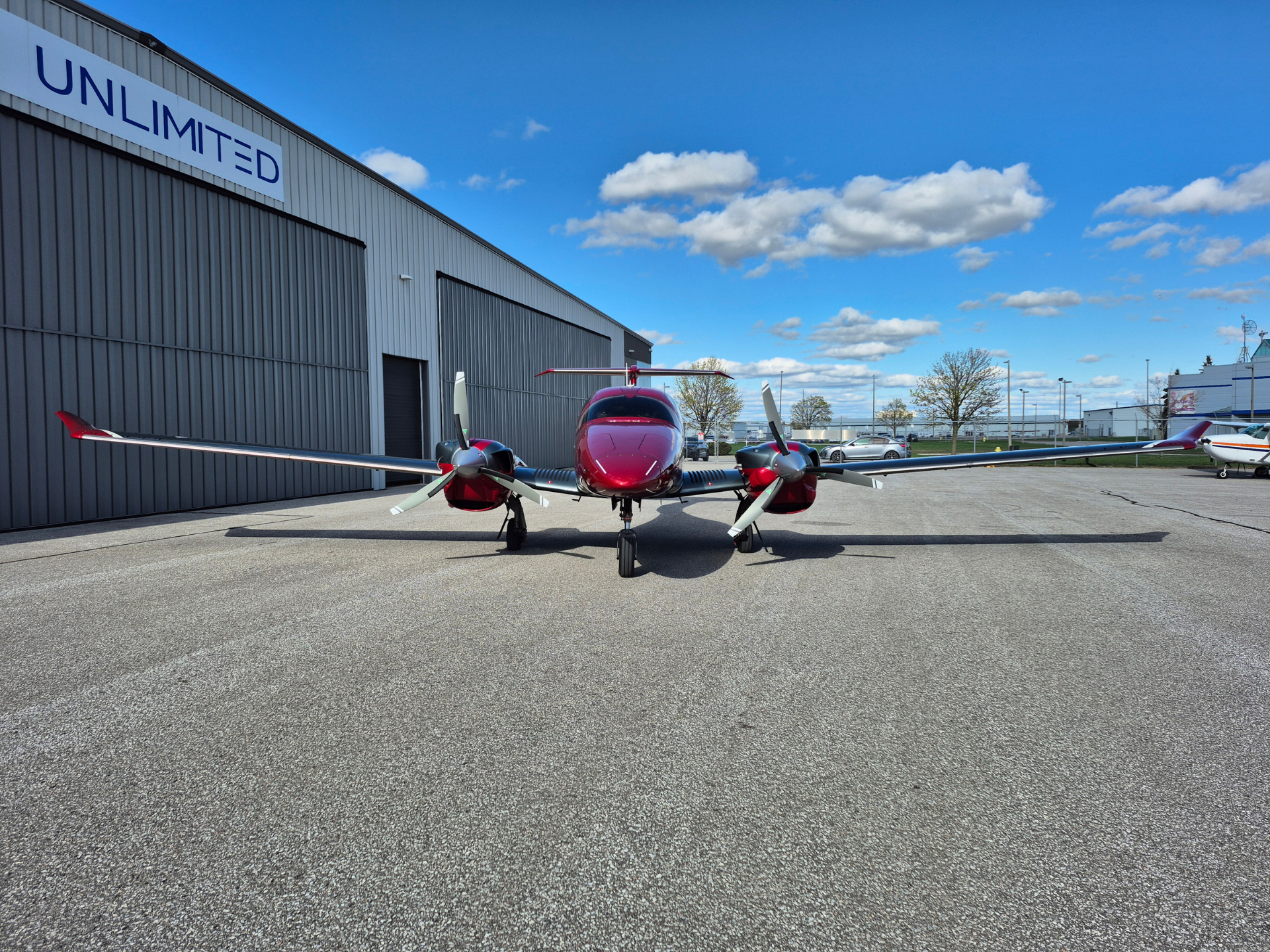 Front view of a red twin-engine airplane parked on a tarmac in front of a gray hangar with a large 'UNLIMITED' sign under a blue sky.