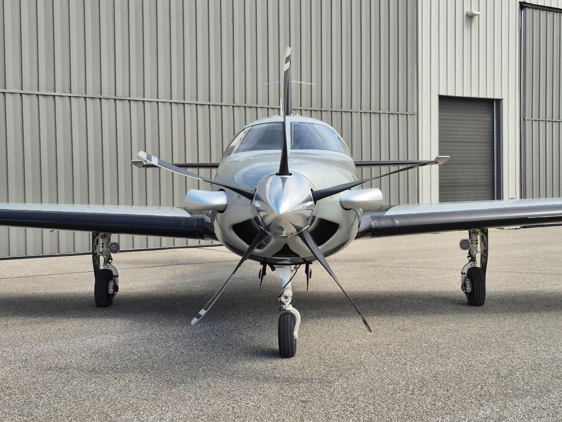 Front view of a small silver propeller airplane facing the camera on a tarmac in front of a corrugated metal hangar.