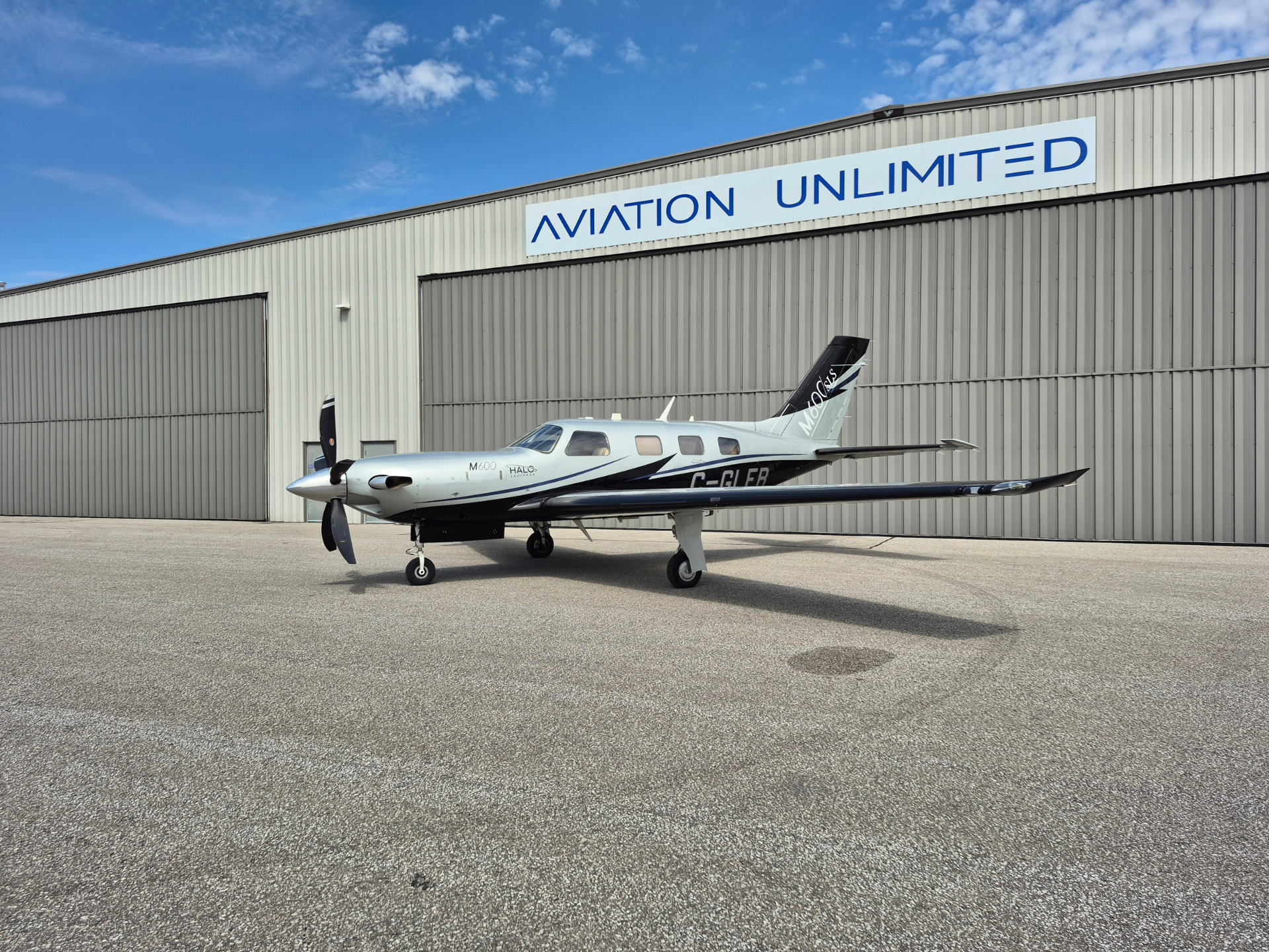 Small silver propeller airplane parked on tarmac in front of a large gray hangar with an 'AVIATION UNLIMITED' sign under a blue sky, clouds.