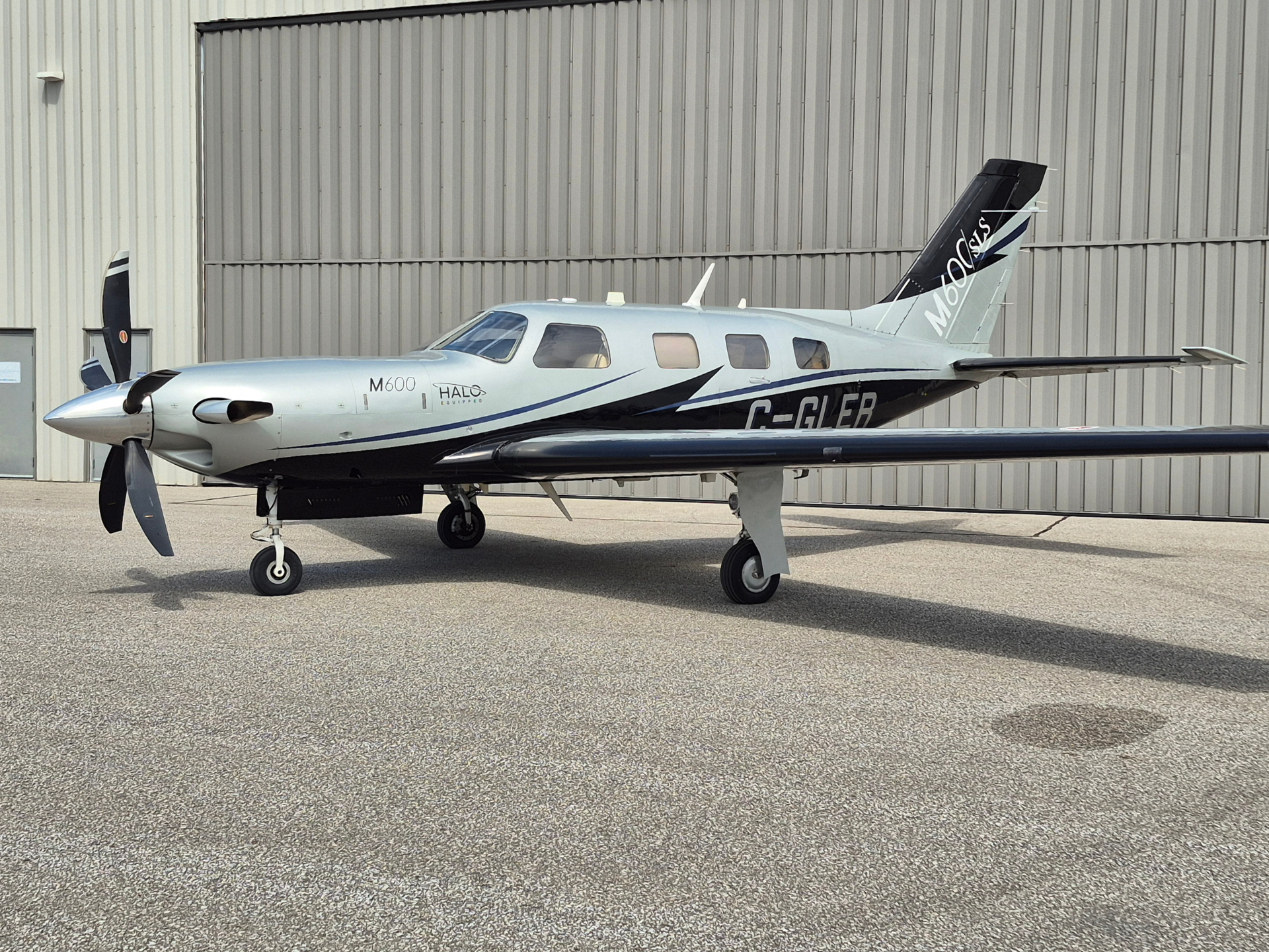 Silver twin-engine propeller aircraft parked on the tarmac beside a large beige hangar, with 'M600' markings on the fuselage and tail.