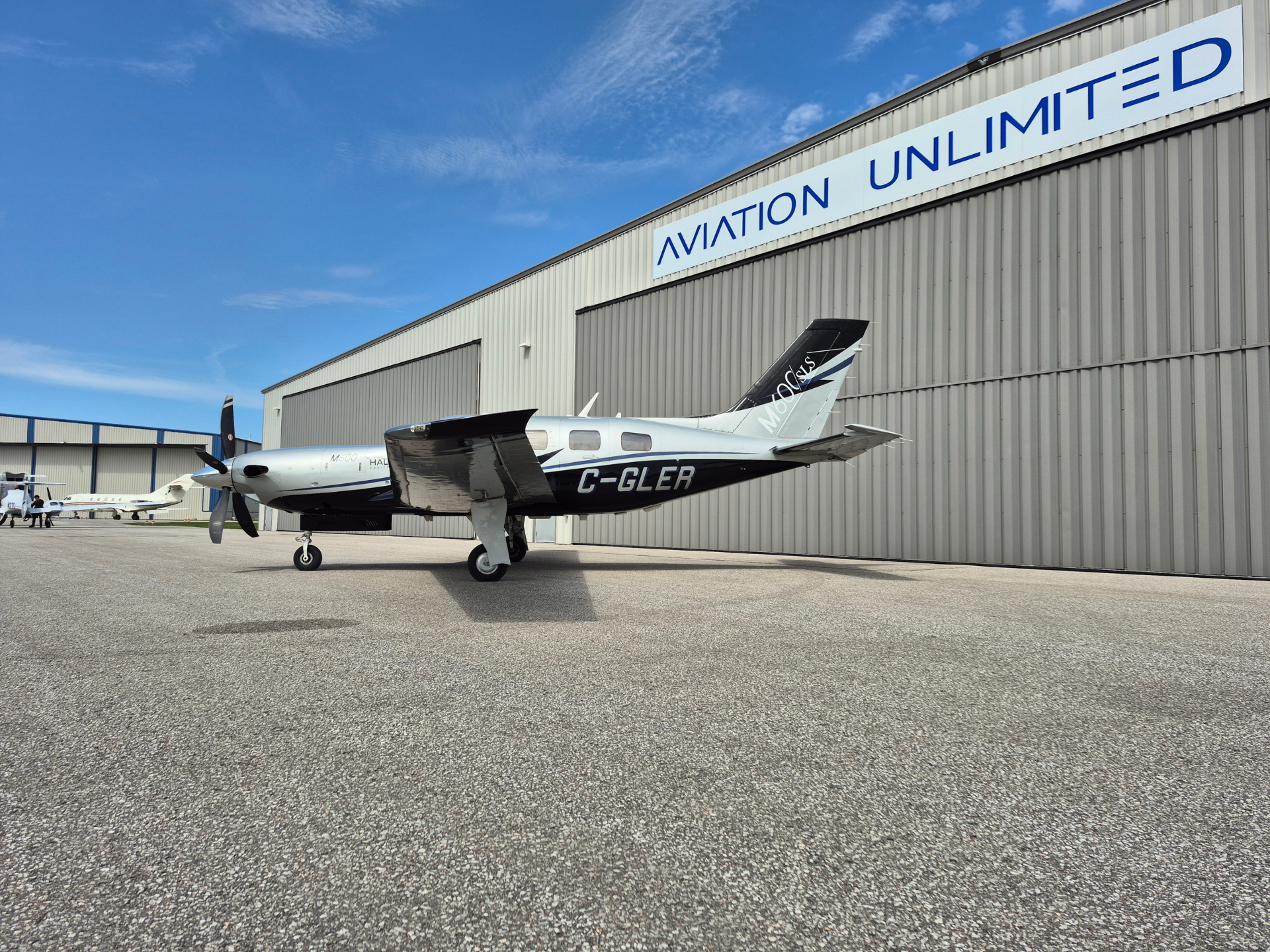 Single-engine propeller aircraft on the tarmac in front of a large corrugated hangar with an 'AVIATION UNLIMITED' sign above.