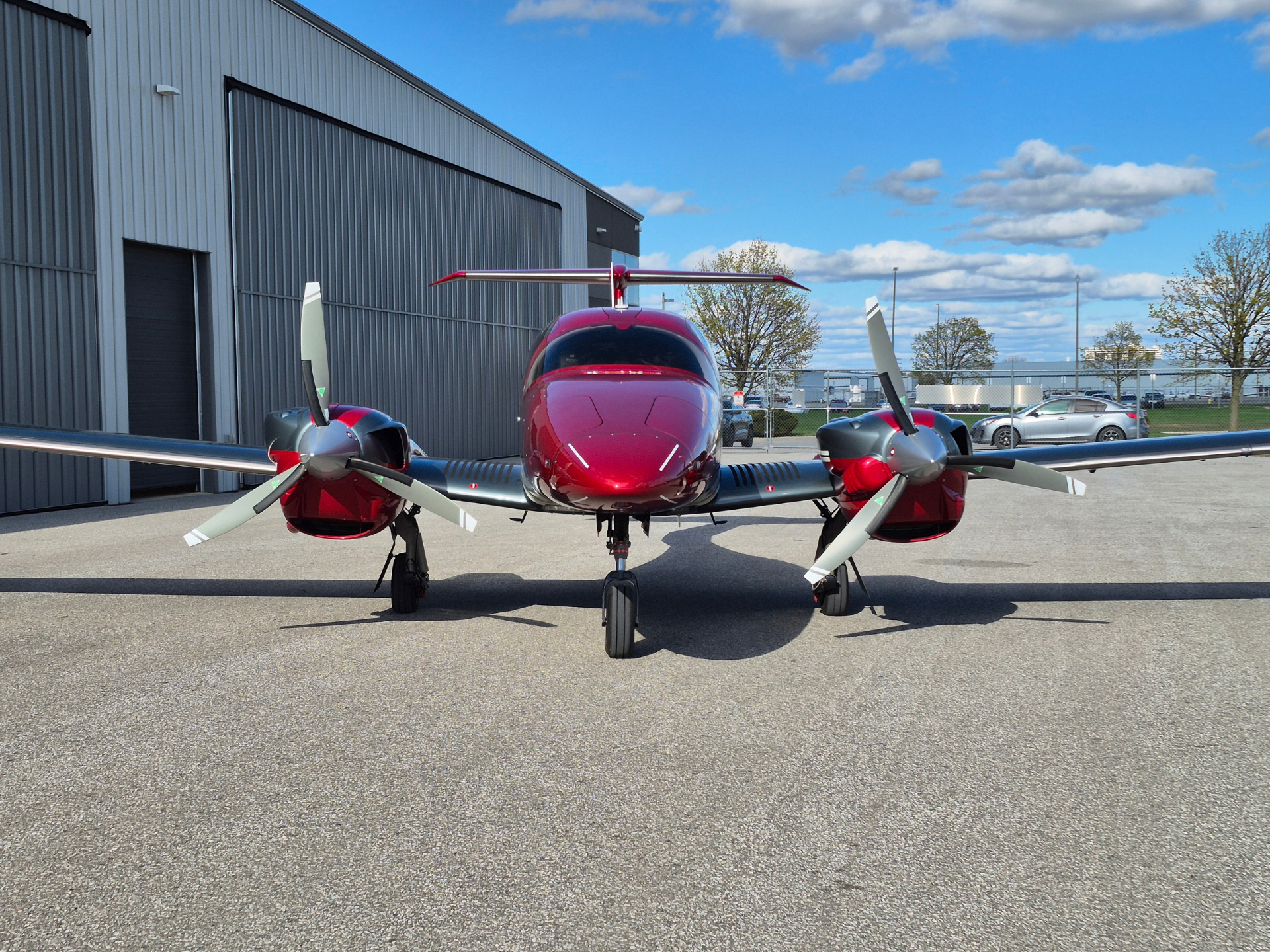 Front view of a red twin-engine propeller airplane parked on a tarmac beside a gray hangar under a blue sky with clouds.