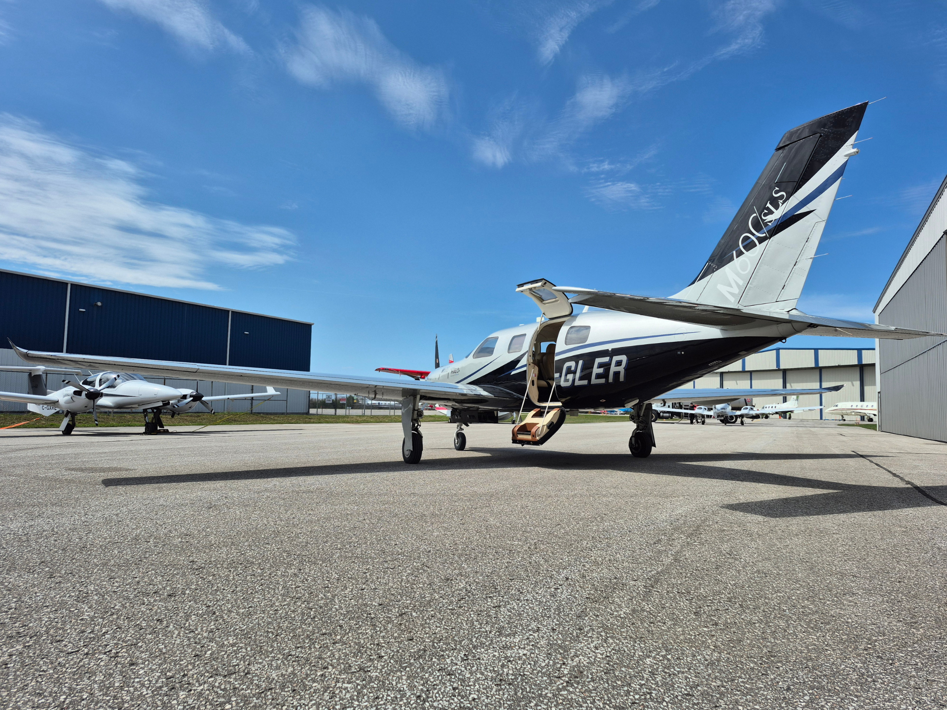 Small private jet parked on a sunlit airport tarmac with an open door and stairs, hangars in the background.