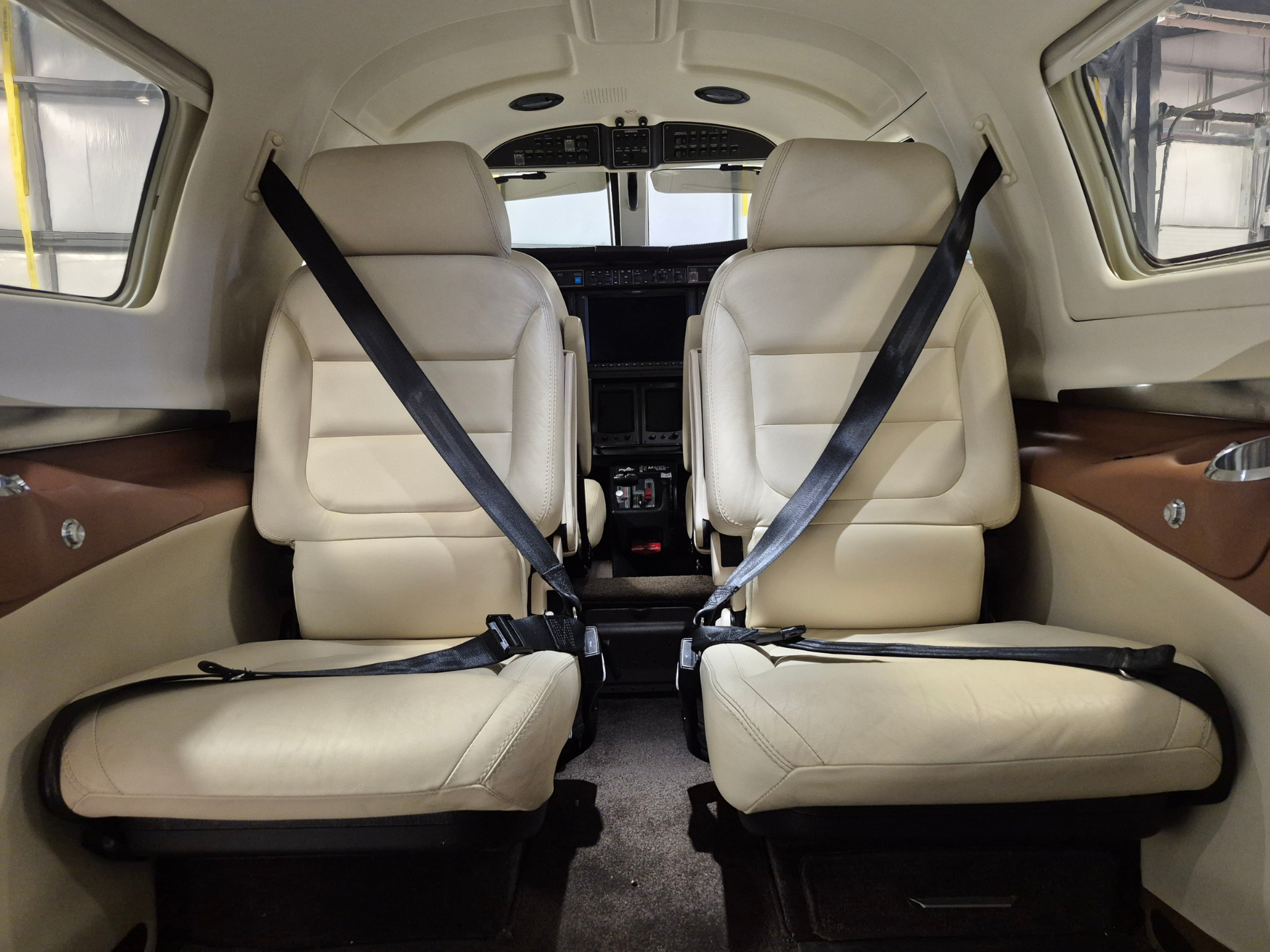 Interior of a light beige airplane cabin with two captain chairs and attached seat belts, cockpit instruments visible ahead.