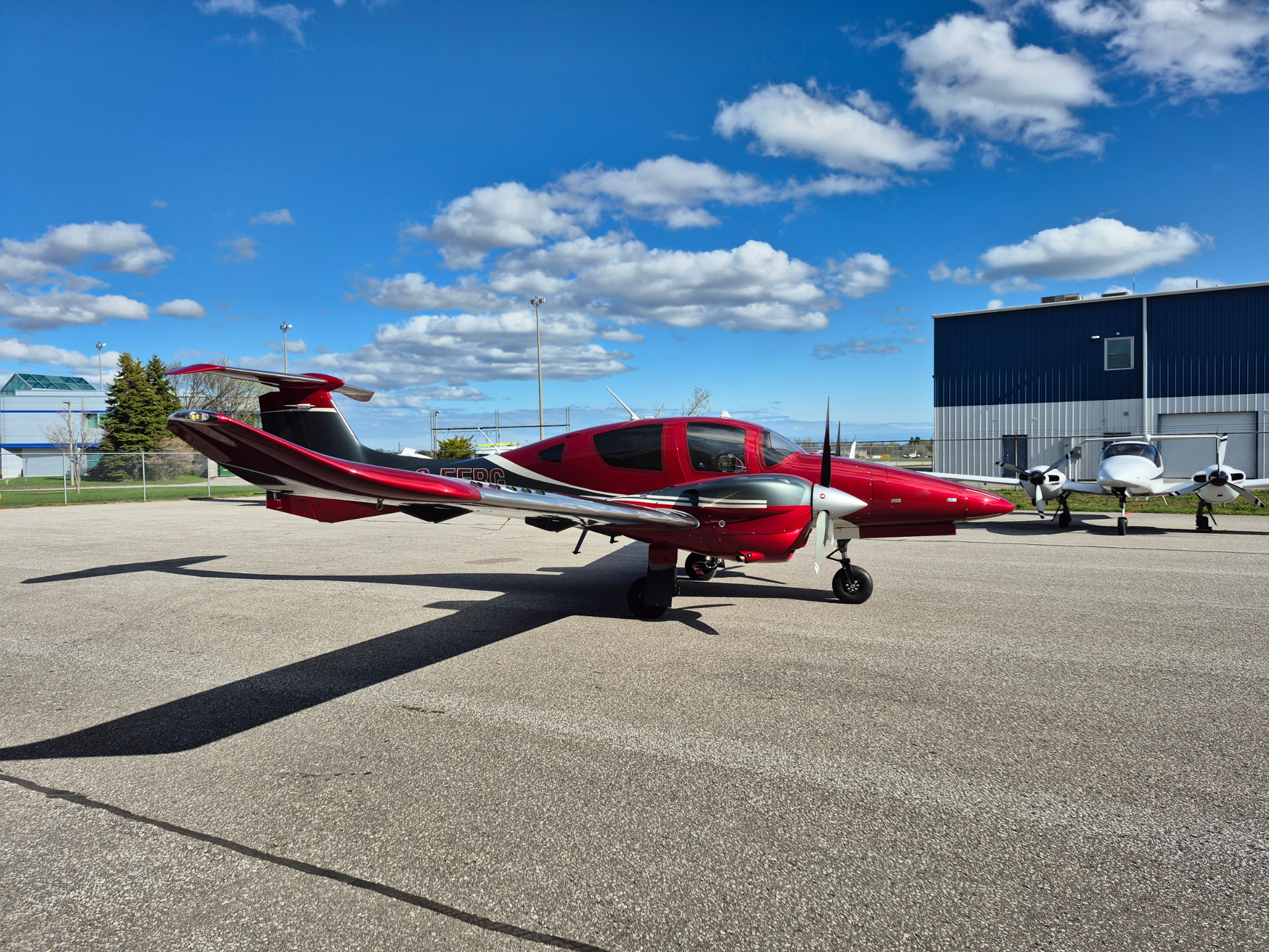 Red small aircraft on a sunlit tarmac with a blue hangar and white clouds in the bright sky.
