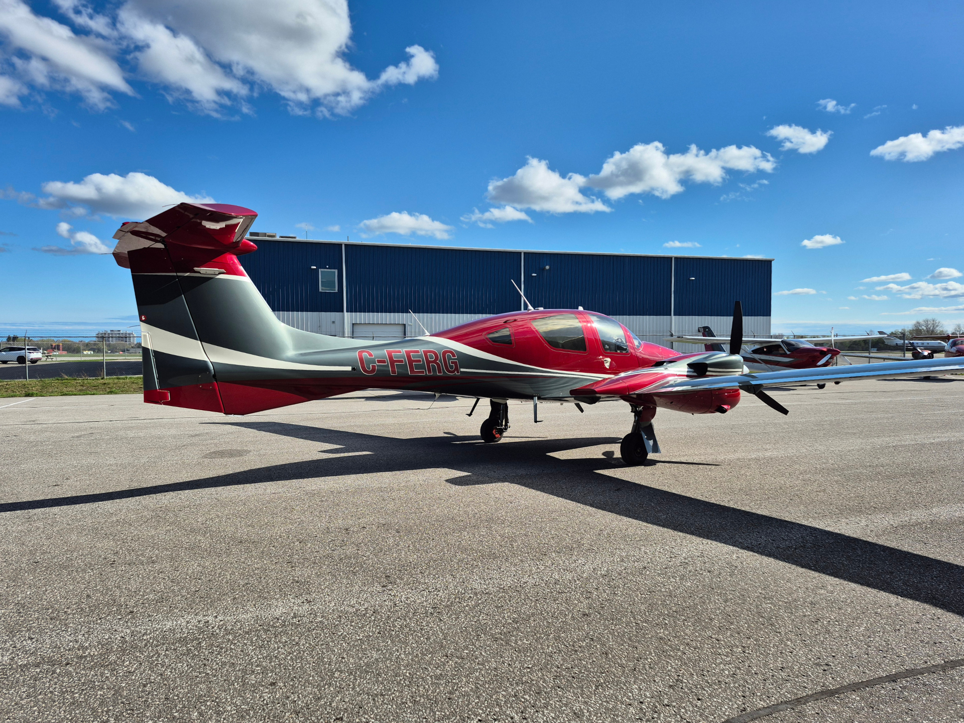 Small red and gray single-engine airplane parked on a tarmac in front of a blue hangar under a bright sky with clouds, tail number C-FERG