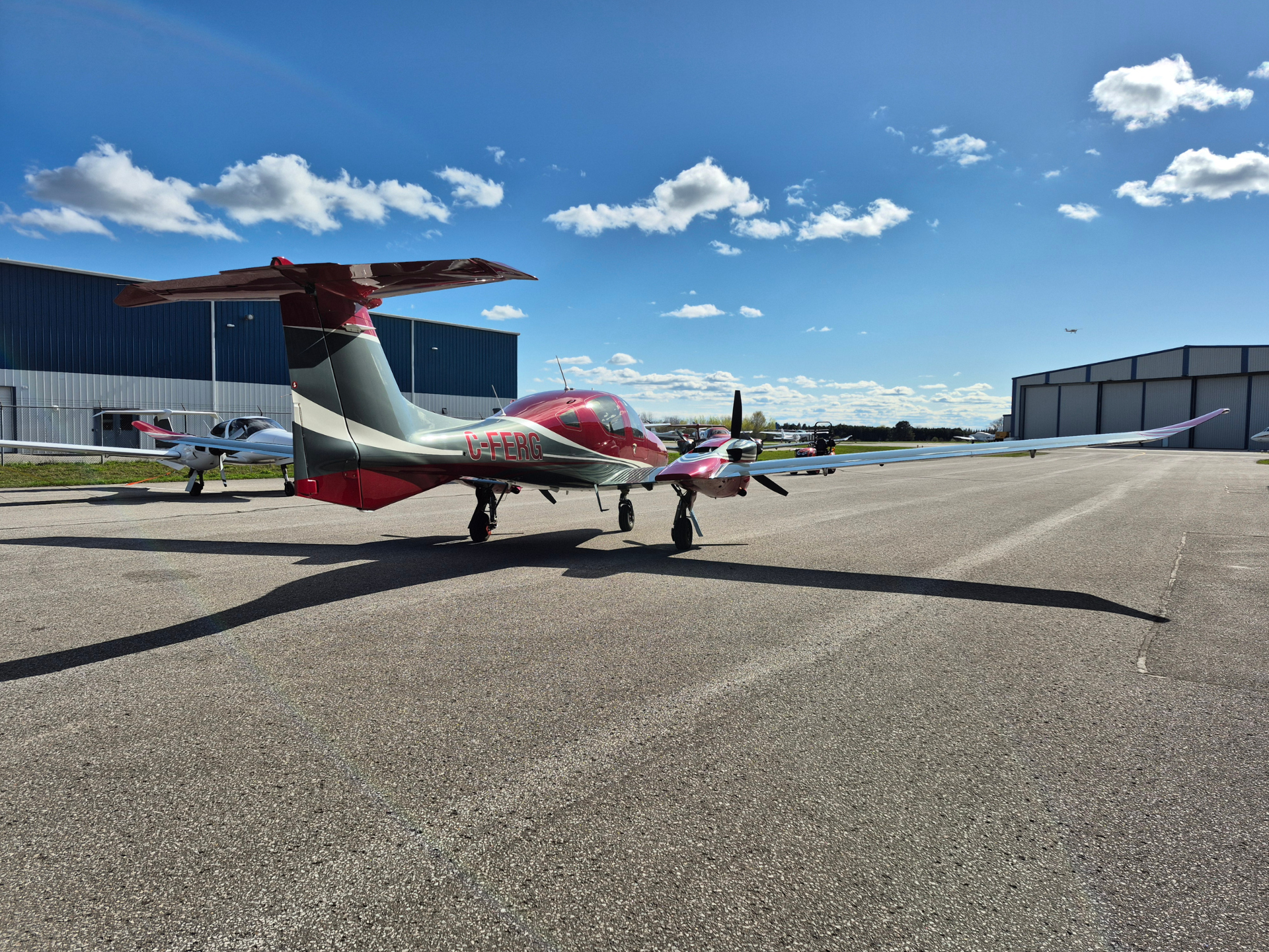 Red and white light aircraft with tail number C-FERG parked on a sunlit tarmac beside blue hangars under a clear blue sky.
