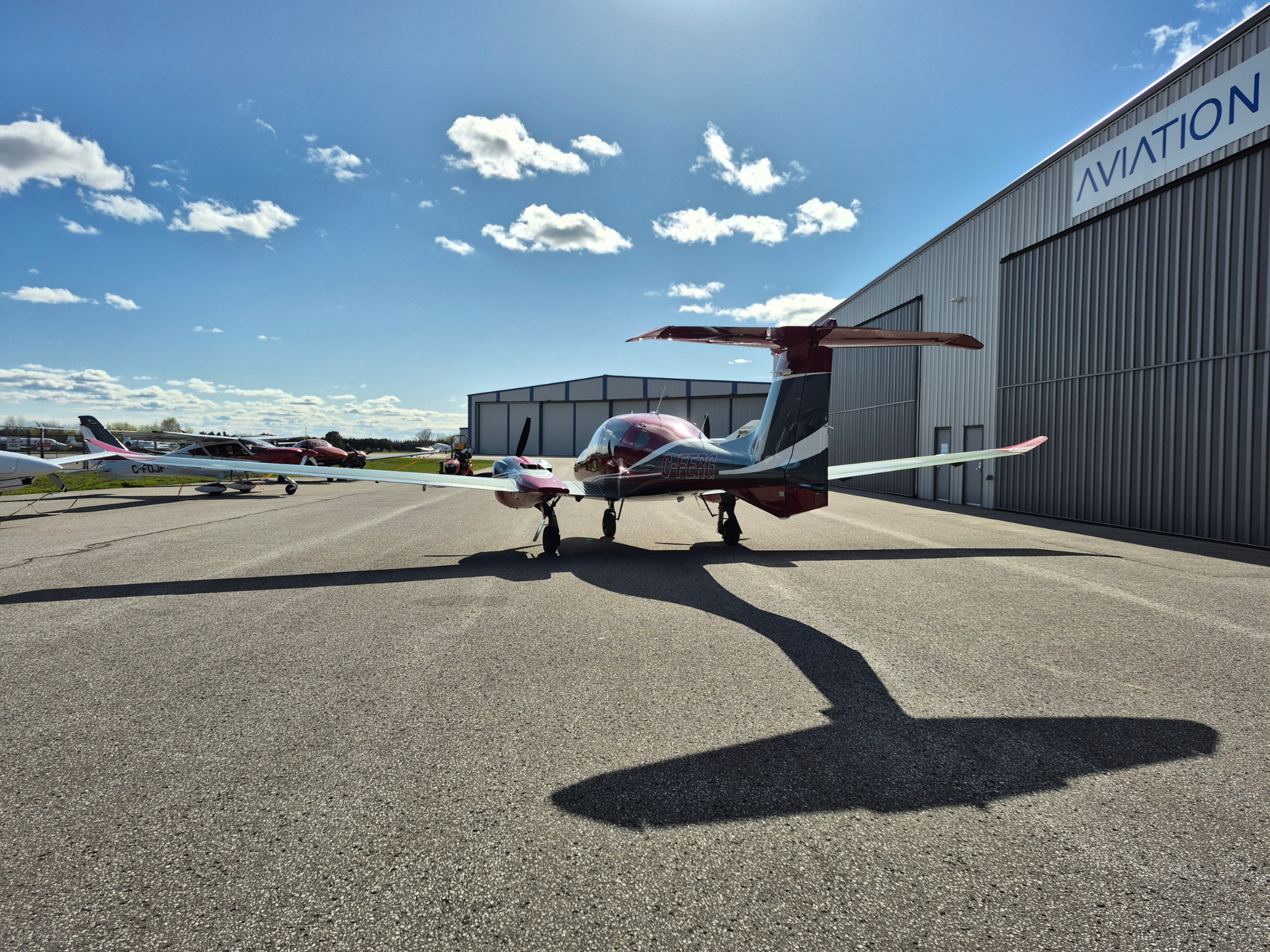 Small red-and-white light aircraft parked on a tarmac beside a large aviation hangar under a blue sky with scattered clouds