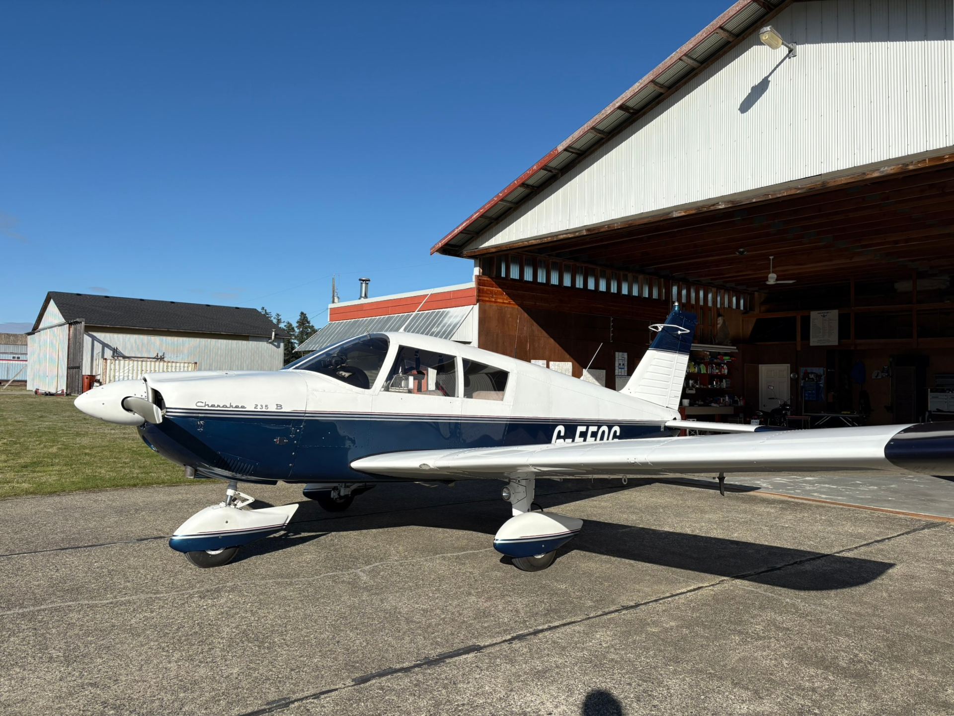 White and blue Cessna Cherokee 235B aircraft parked on the tarmac outside a hangar against a clear blue sky. (informative)