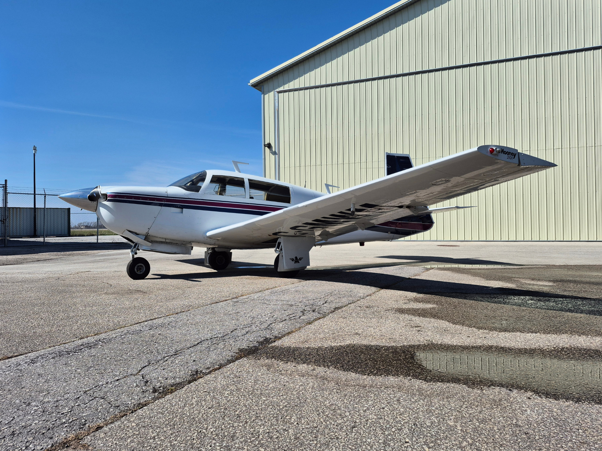 Small white propeller airplane with a red and blue stripe parked on a cracked tarmac beside a pale yellow hangar under a clear blue sky.