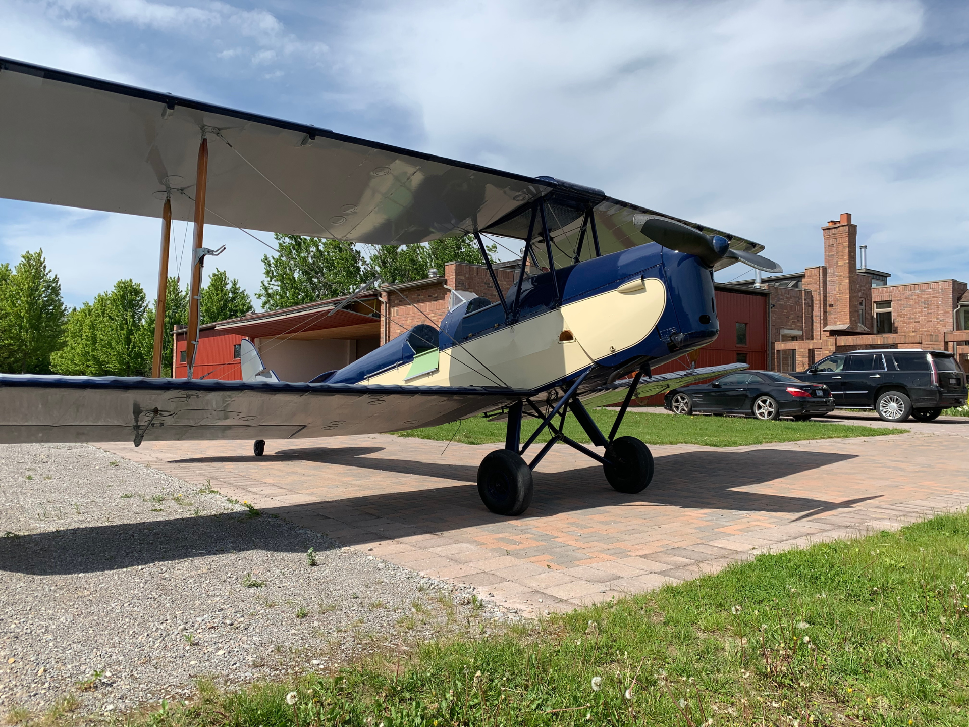Small vintage biplane on a sunlit tarmac with brick buildings and parked cars in the background.