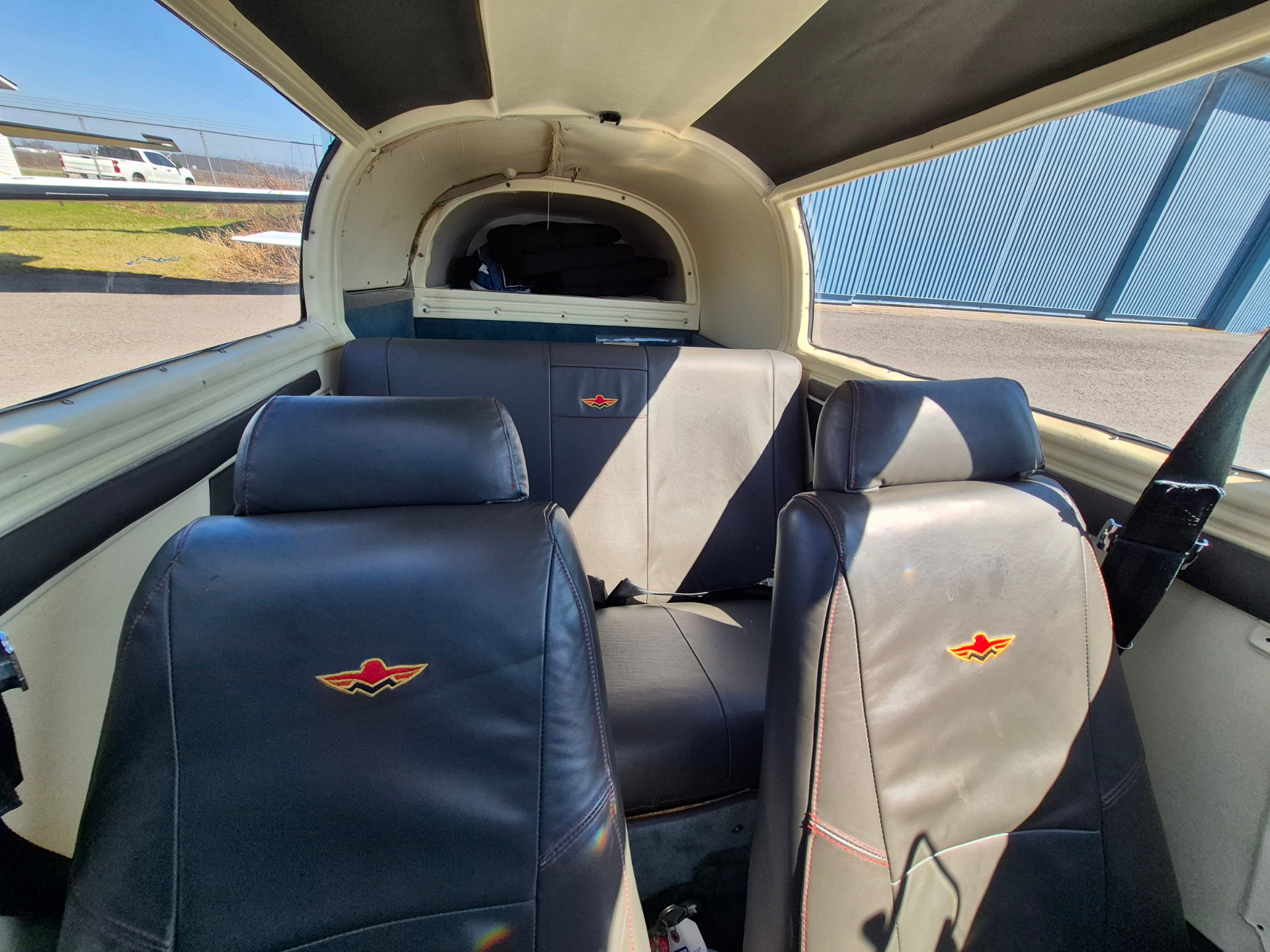 Interior of a small airplane cabin with dark leather seats and a stack of belongings in the rear, side windows revealing tarmac and hangar.