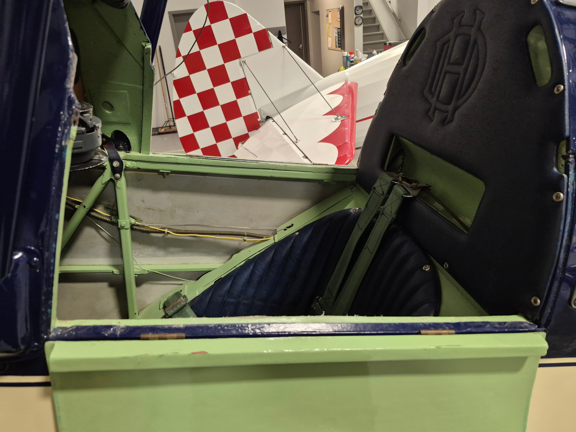 View into a vintage aircraft cockpit with green metal frame, blue seat padding, and a red-and-white checkered tail surface visible through the doorway.