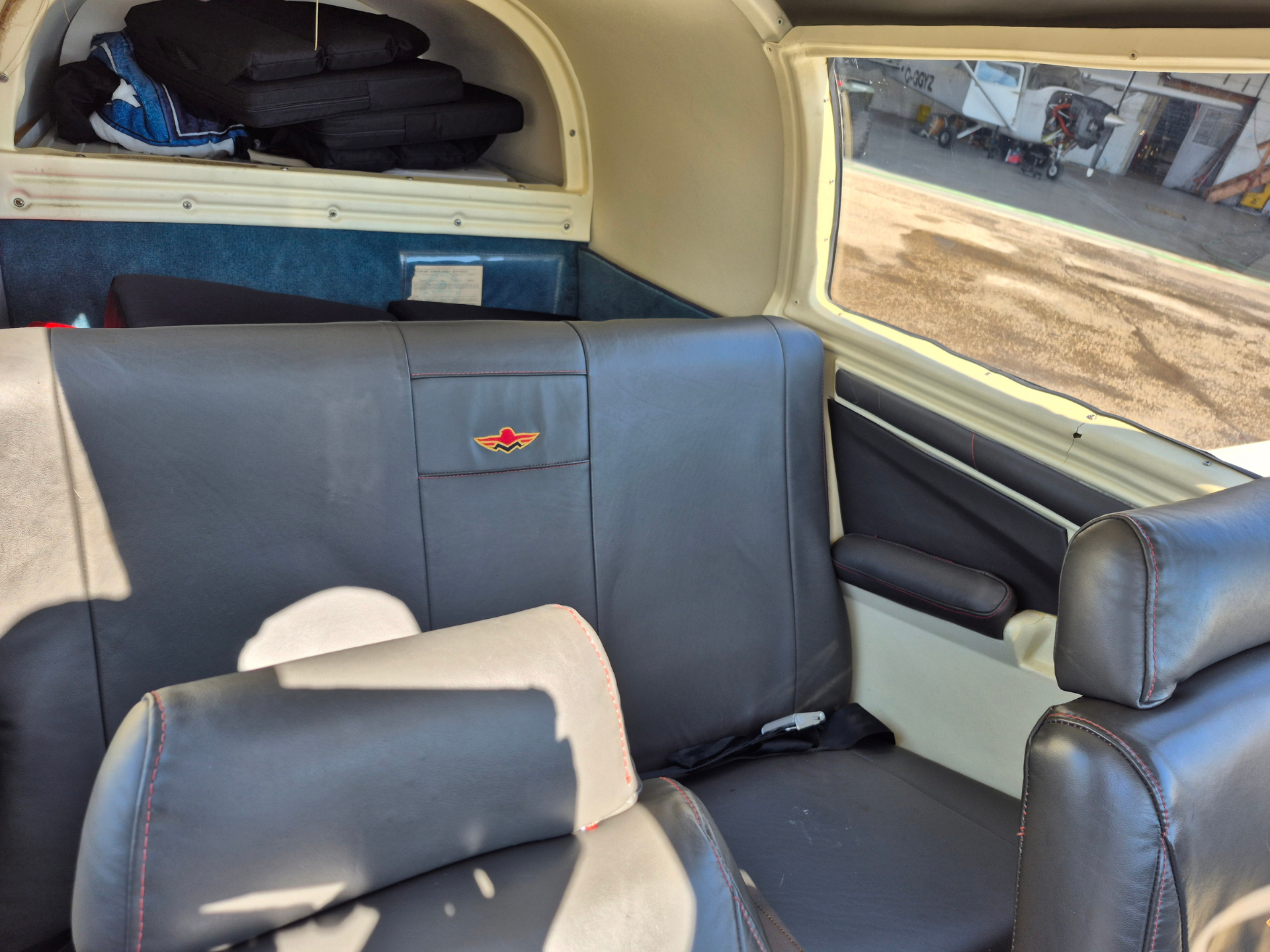 Interior of a small airplane cabin with blue leather seats and red stitching, overhead baggage area with folded items, window on the right side