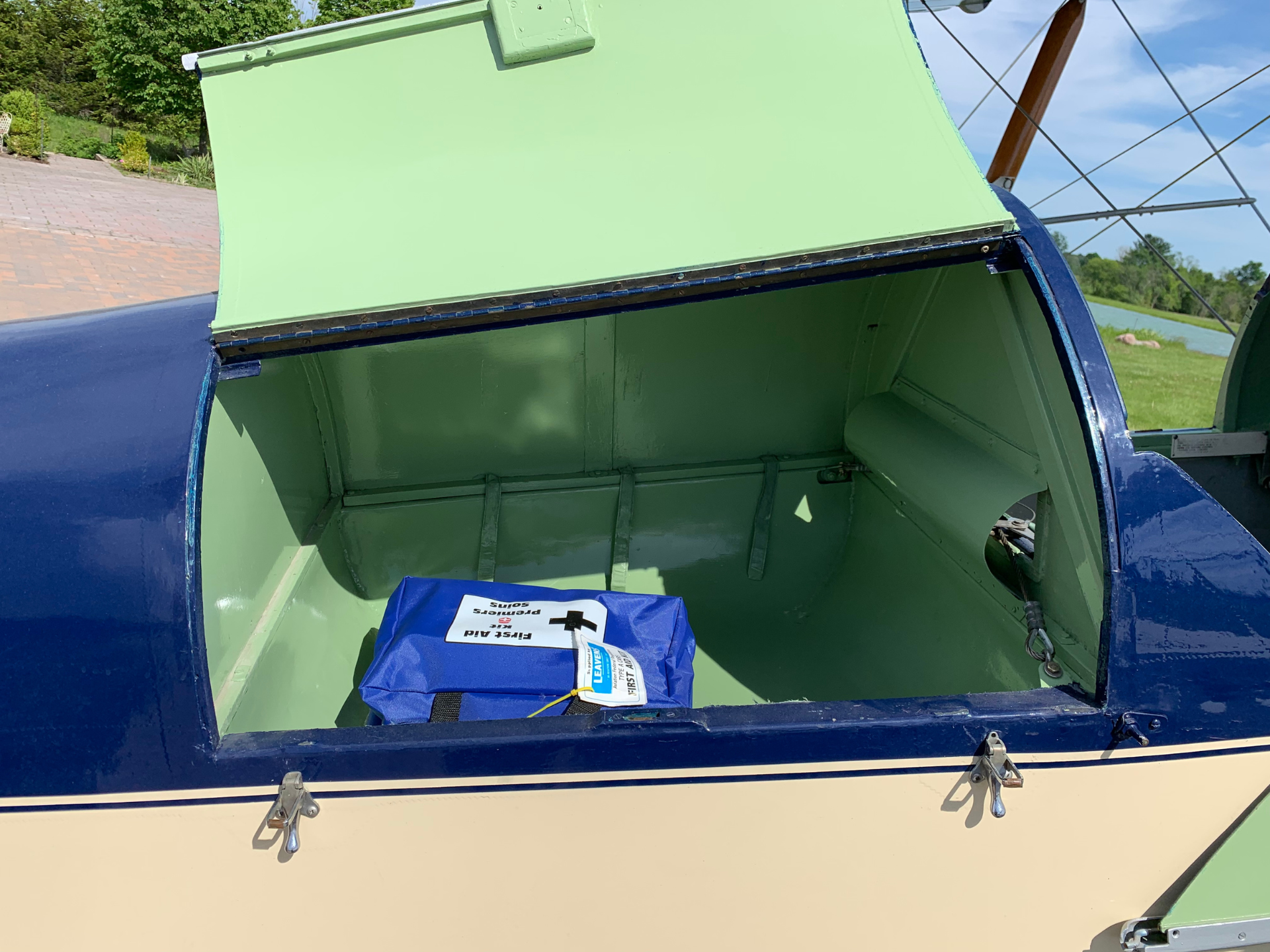 Open aircraft hatch revealing green interior and a blue first aid kit labeled 'First Aid Supplies' inside, with blue exterior and outdoor scenery in the background.