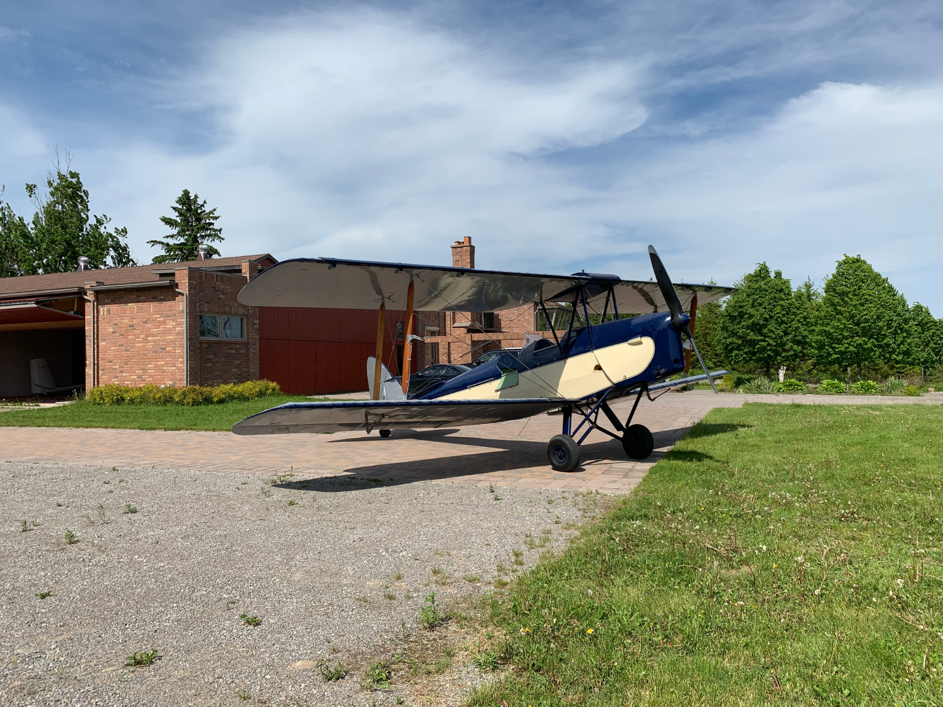 Small vintage biplane parked on a brick driveway in front of a brick house under a blue sky with scattered clouds.