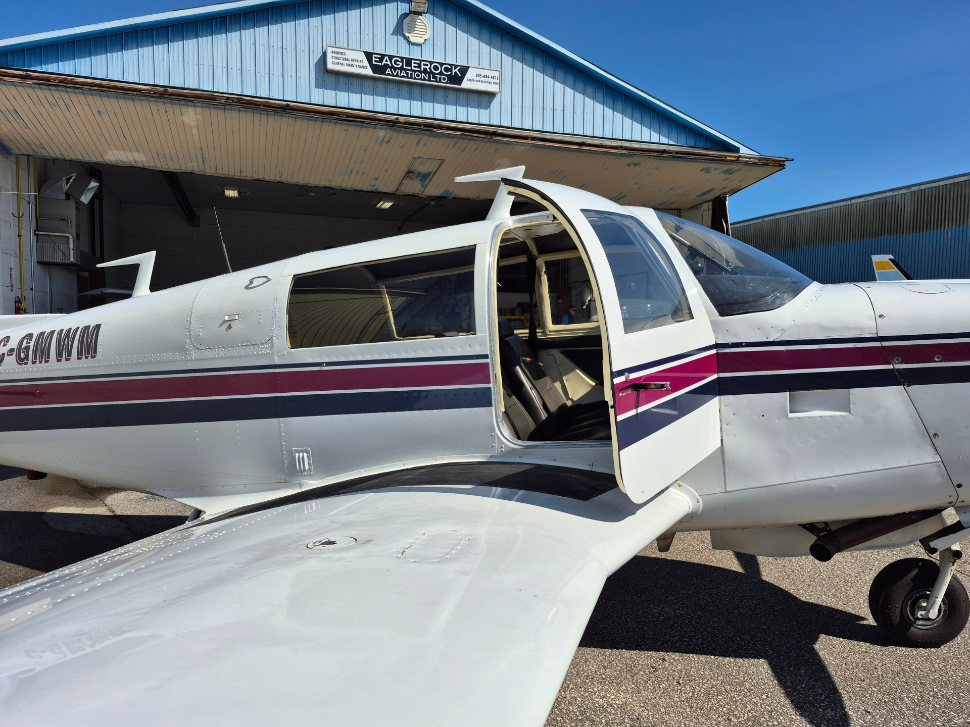 Small white single-engine airplane with open door parked on tarmac beside a blue hangar; cockpit and passenger seats visible inside.