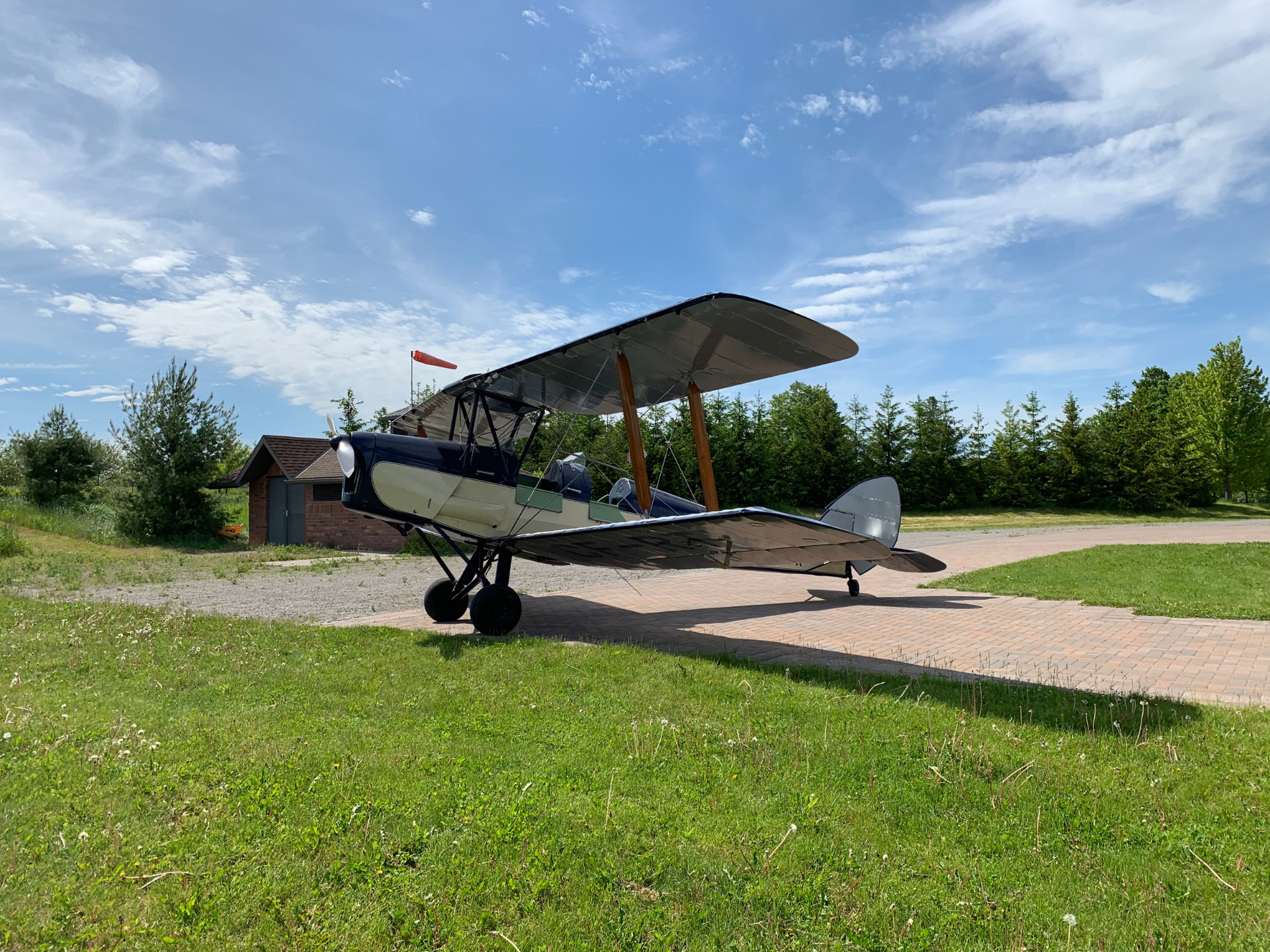 Vintage biplane parked on a brick path beside a small brick hangar, with green grass and trees under a bright blue sky.
