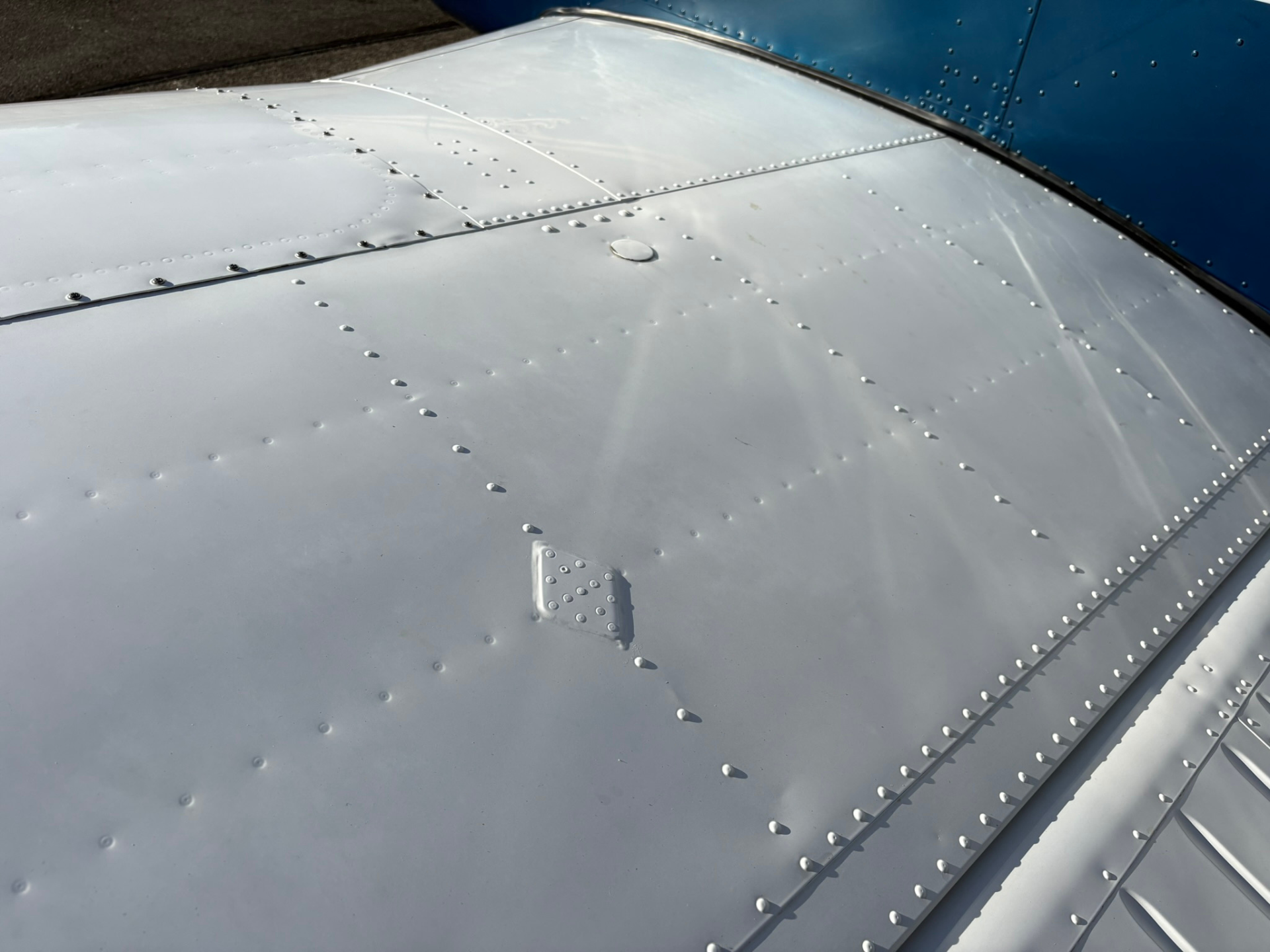 Close-up of a light gray aircraft wing surface showing rivets and panel seams, with a blue fuselage edge in the background