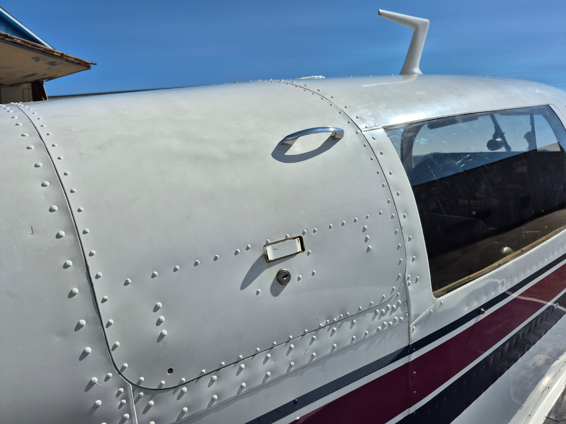 Close-up of a small aircraft fuselage: riveted silver metal with cockpit window and door handle, under a clear blue sky