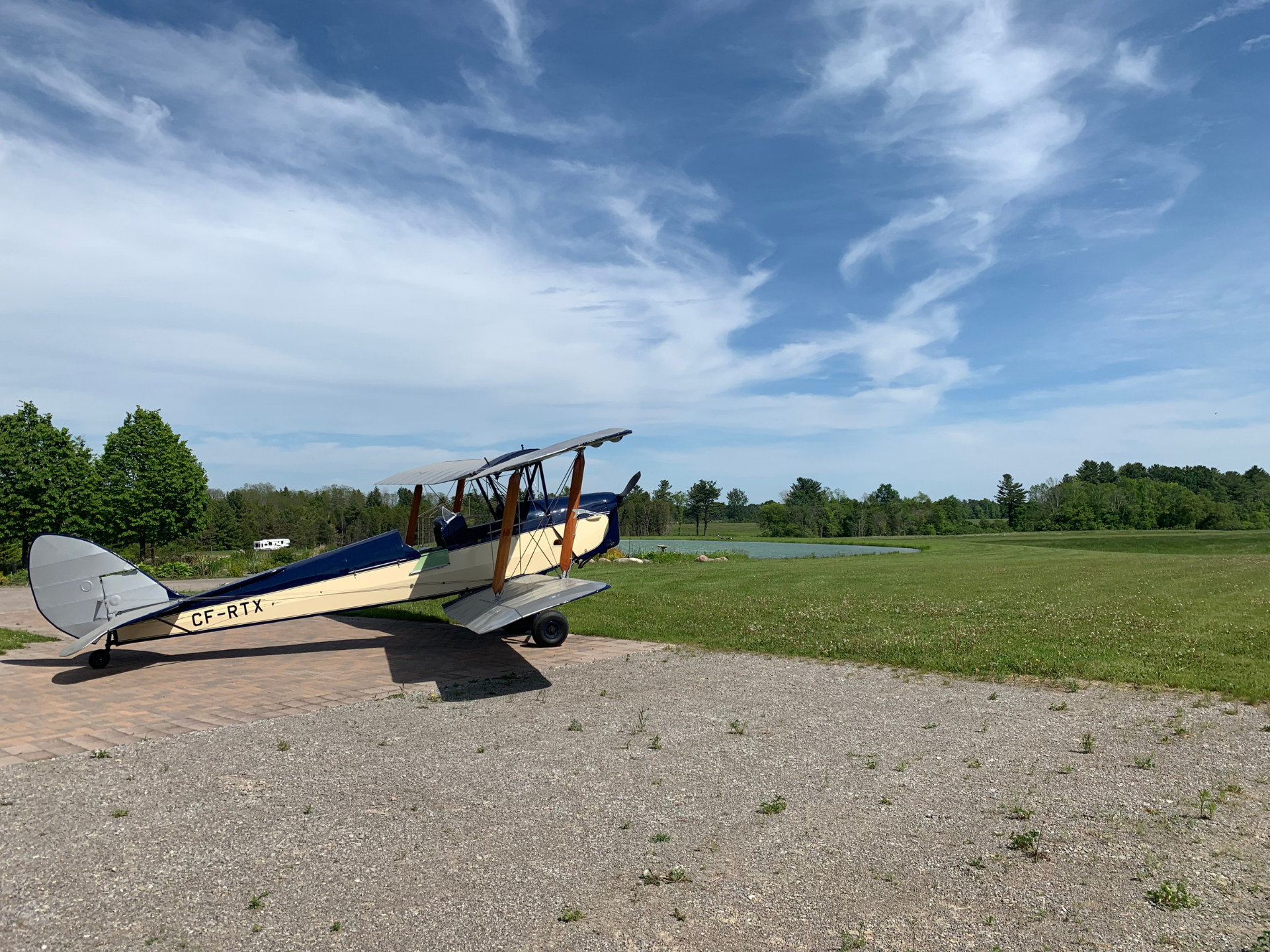 Vintage biplane parked on a paved path beside a grassy field under a blue, cloudy sky; CF-RTX visible on the fuselage.