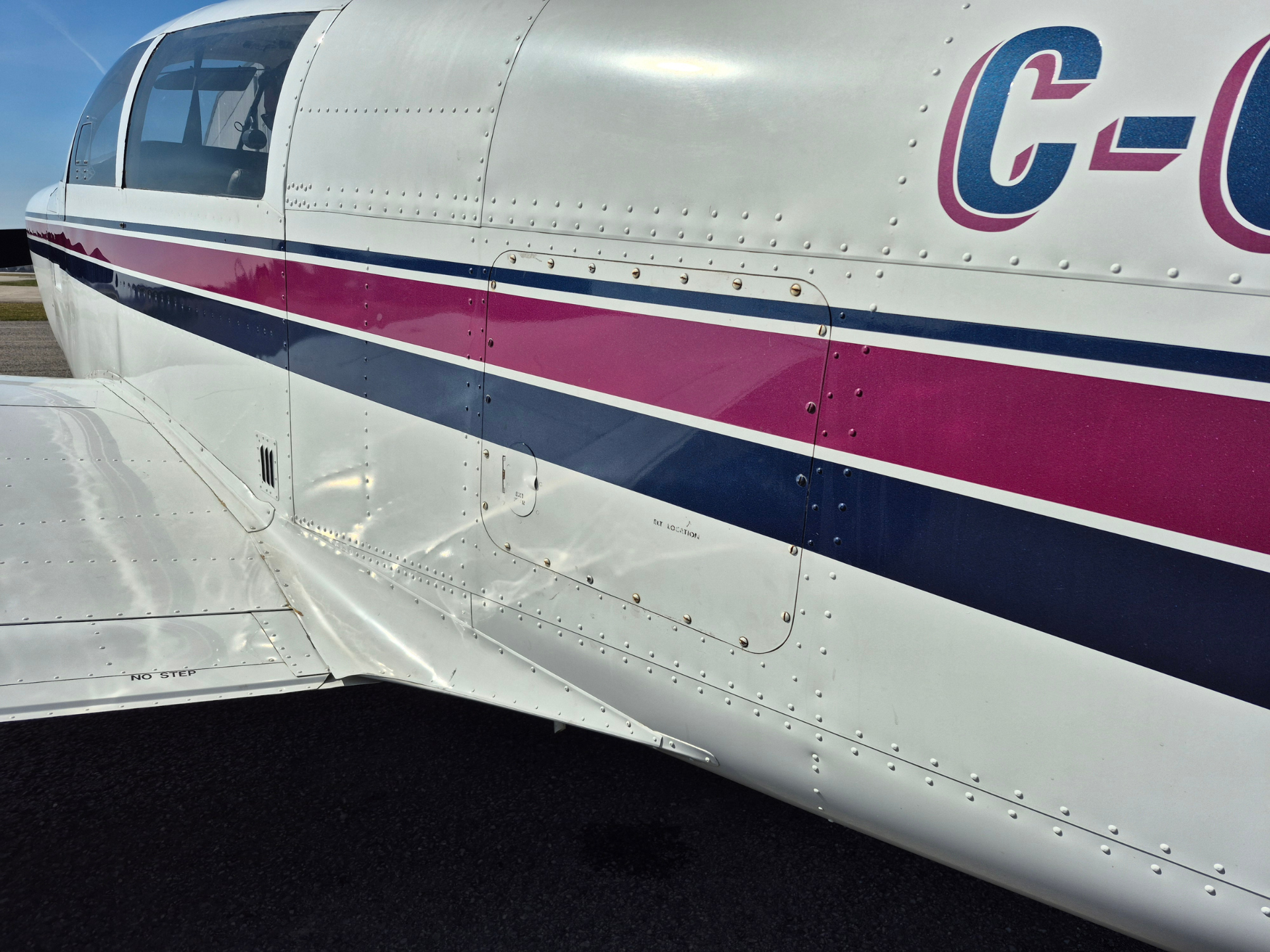 Side view of a small white airplane fuselage with pink, white and blue stripes and riveted metal panels near the cockpit window