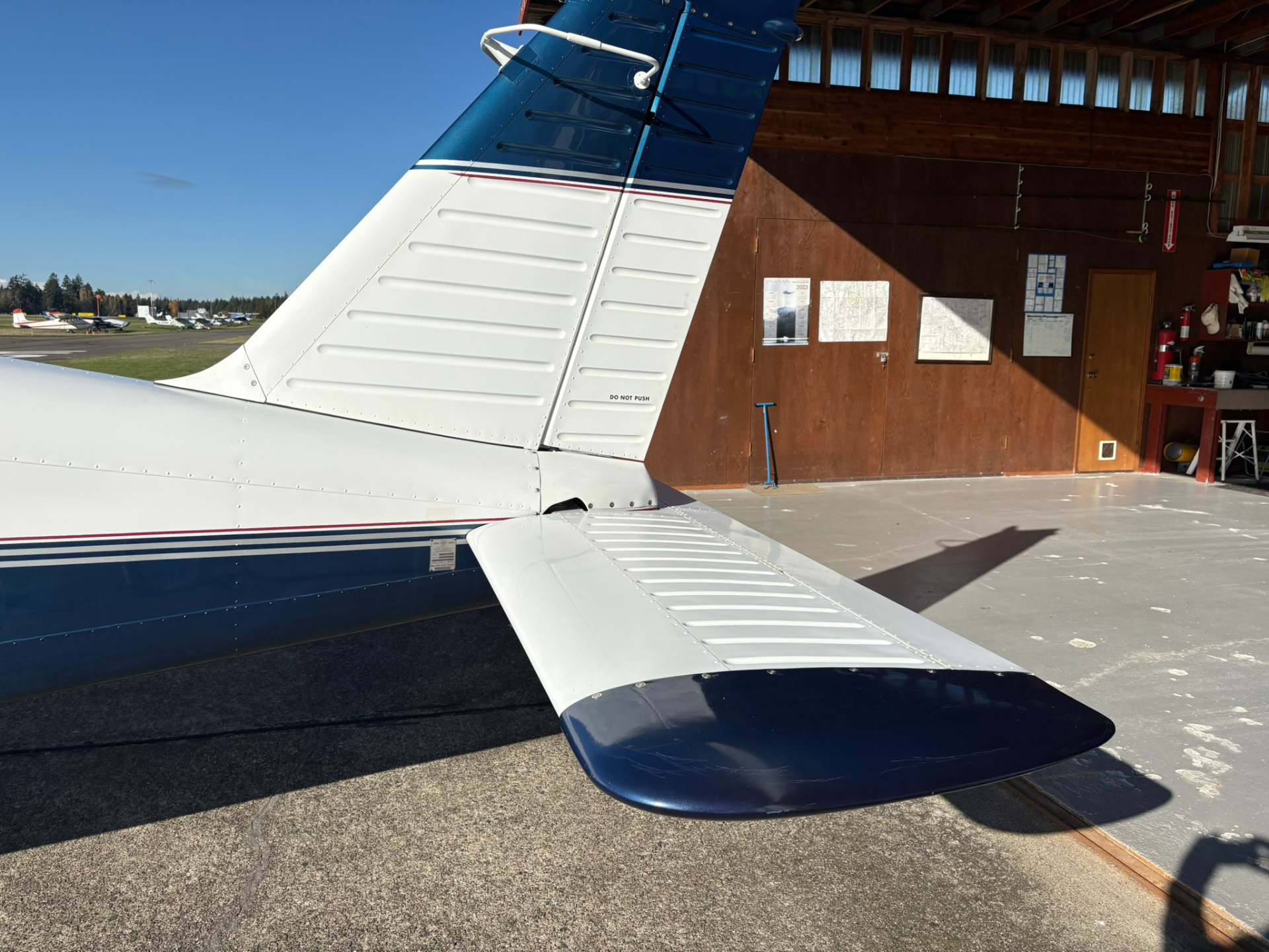 Wing and tail section of a small propeller airplane inside a hangar, with other planes visible outside and blue sky beyond.
