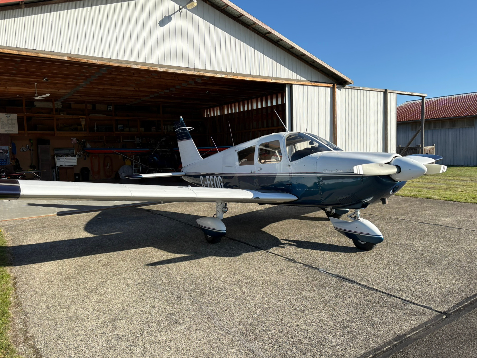 Small white and blue single-engine airplane parked on a tarmac in front of a hangar under a clear blue sky.