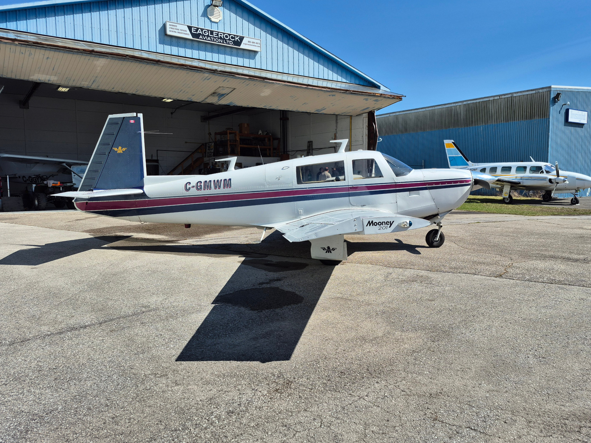 Mooney 201 light aircraft with maroon stripe parked on the tarmac in front of a blue hangar, another plane visible in the background