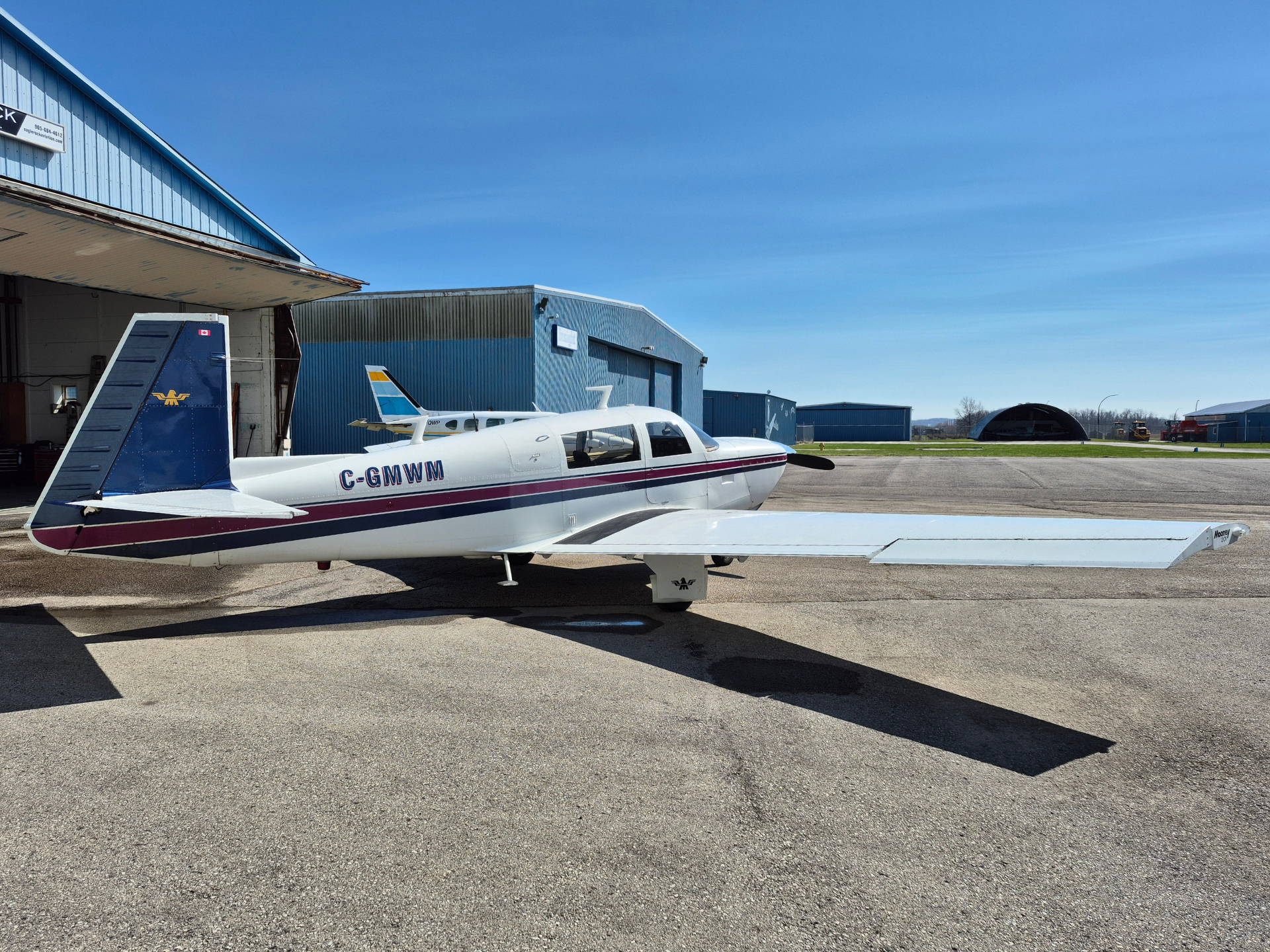 Small white single-engine airplane with blue and maroon stripes parked on the tarmac beside blue hangars.