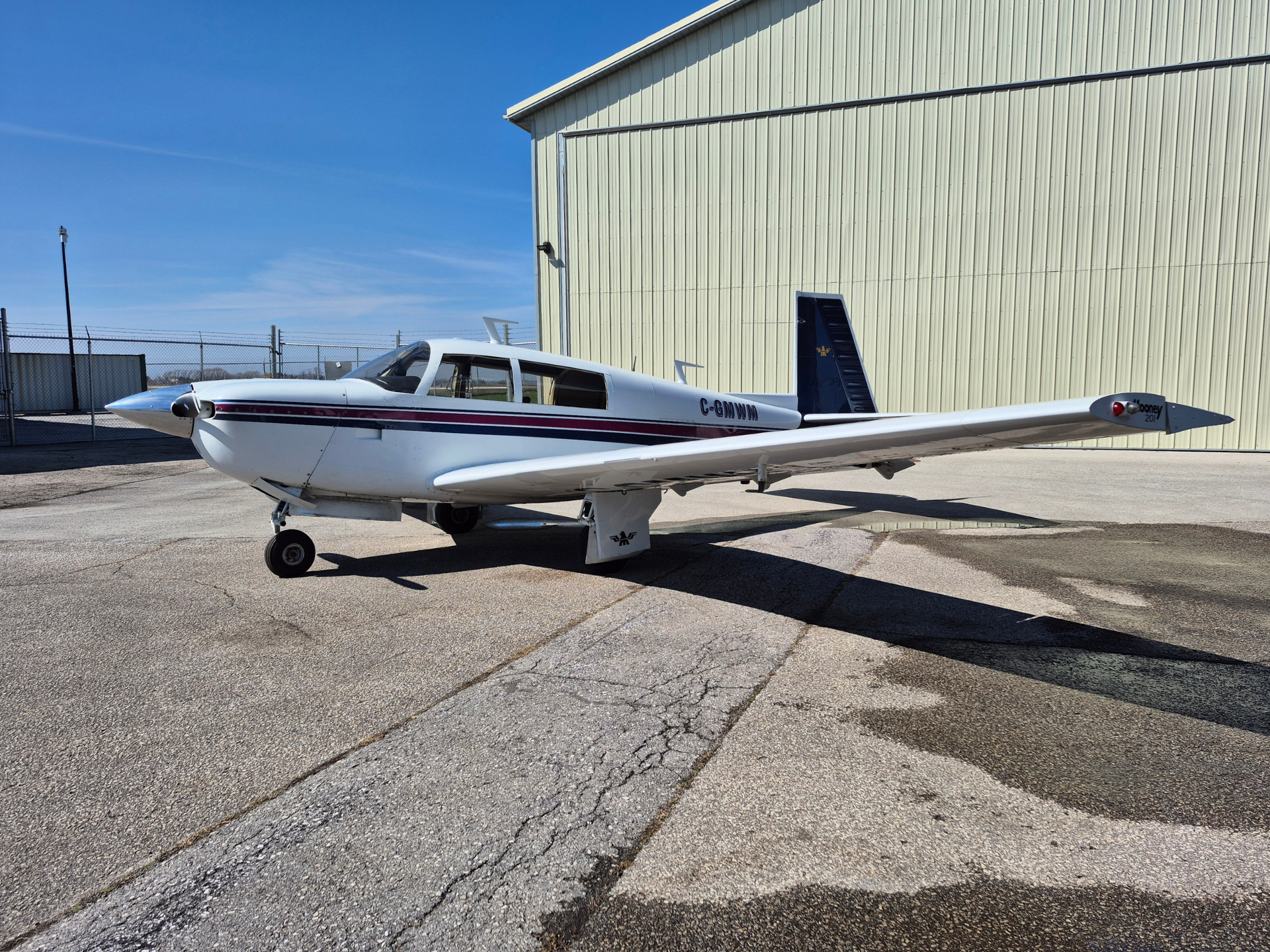 White single-engine propeller airplane parked on a cracked apron beside a large beige hangar under a clear blue sky.