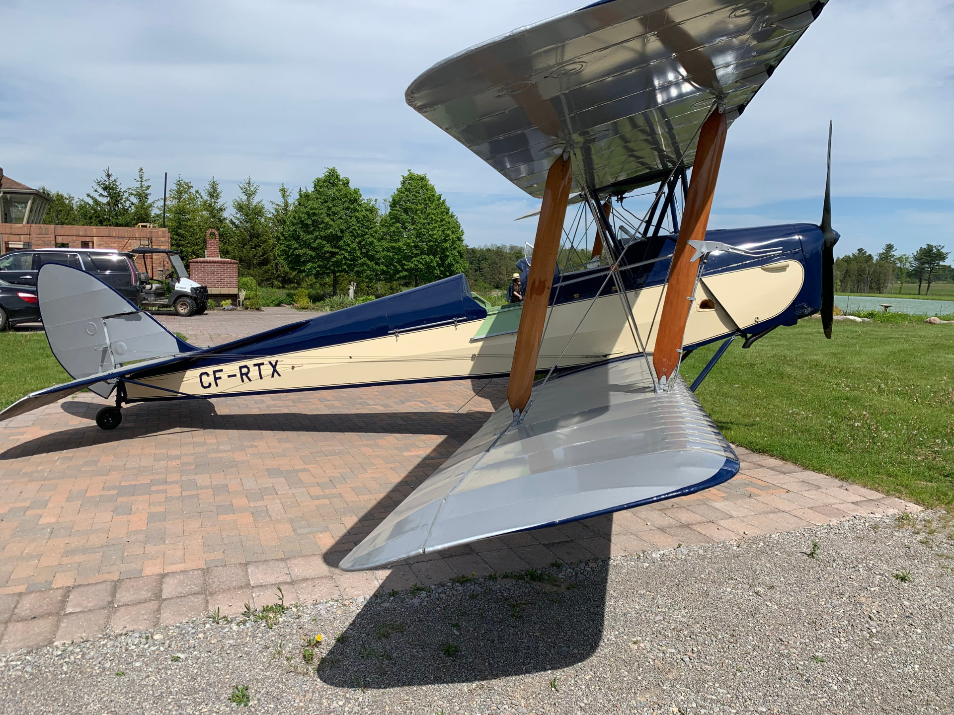 Vintage biplane with blue and cream fuselage parked on a brick driveway; wooden wing struts and a propeller visible at the nose, registration CF-RTX.