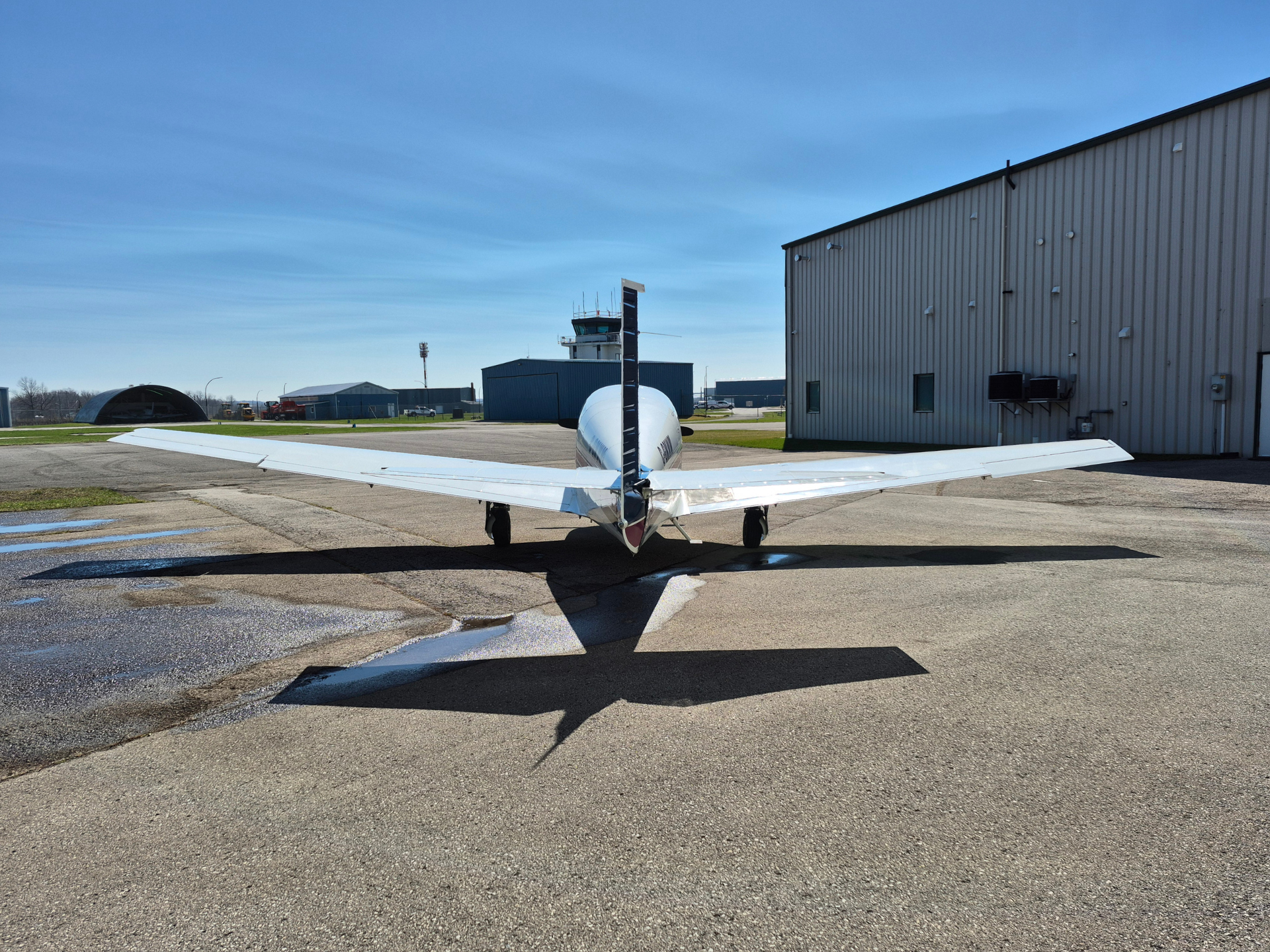 Small white propeller airplane on a tarmac near a large gray hangar under a bright blue sky, airport buildings in the background.